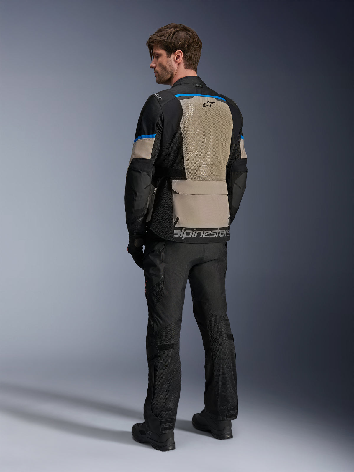 Kurtka Alpinestars Andes Air Drystar®, czarna, szara Laurel Gray i jasnoniebieska, widok z tyłu ukazujący duży siatkowy panel wentylacyjny na plecach, logo na dolnej krawędzi, w zestawie z czarnymi spodniami motocyklowymi i butami.
