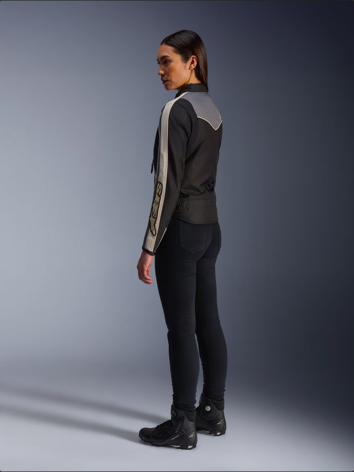 Modelka widziana z boku i z tyłu w kurtce Alpinestars Stella T-Dyno WR w kolorze Black Dark Gray Ecru, z designem inspirowanym stylem vintage, pasami ecru na rękawach i szarym panelem na plecach, zestawiona z czarnymi spodniami i czarnymi butami motocyklowymi.