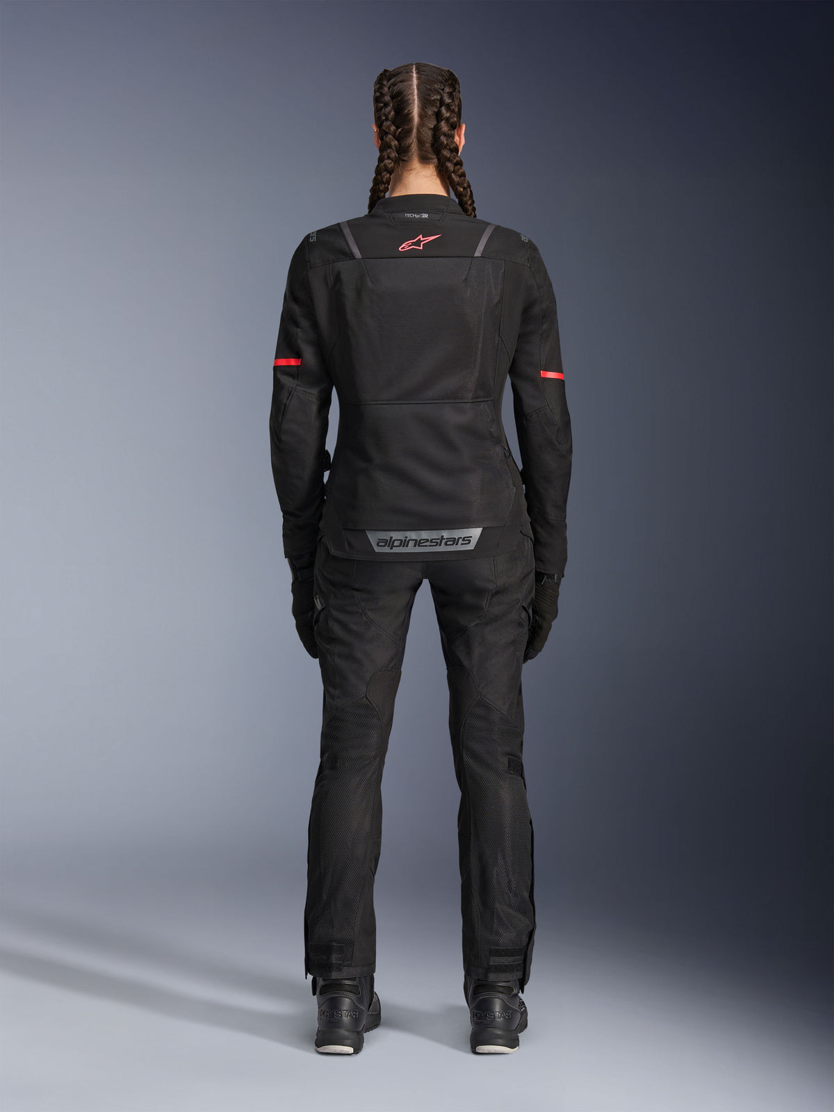 Alpinestars Stella ST-2 Air Jacket, damska kurtka motocyklowa, czarna, widok z tyłu z czerwonym logo na górnej części pleców i szarym oznakowaniem na dole, noszona z dopasowanymi czarnymi spodniami i butami, nowoczesny design turystyczny z panelami z siatki i odblaskowymi detalami