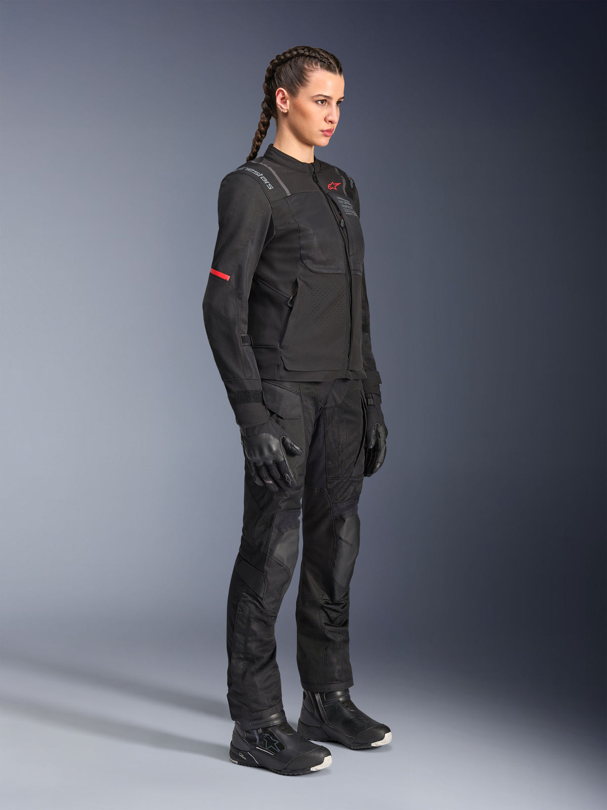Modelka w czarnej kurtce Alpinestars Stella ST-2 Air, w zestawie z dopasowanymi czarnymi spodniami motocyklowymi, rękawicami i butami, siatkowe panele wentylacyjne i subtelne czerwone detale, elegancki nowoczesny strój turystyczny