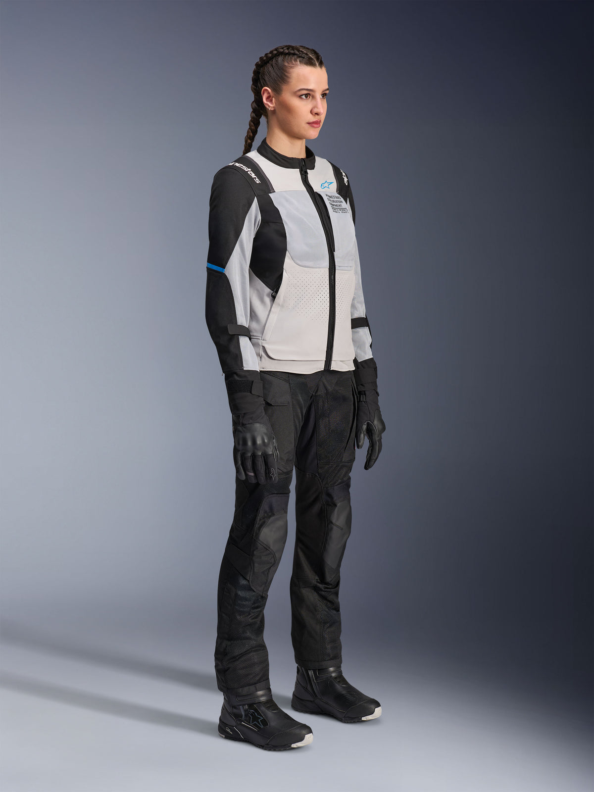 Modelka w kurtce Alpinestars Stella ST-2 Air w kolorze Ice Gray i czarnym, damska techniczna kurtka motocyklowa z panelami z siatki i konstrukcją softshell, w zestawie z czarnymi spodniami i butami adventure touring, z niebieskimi akcentami i nowoczesnym designem