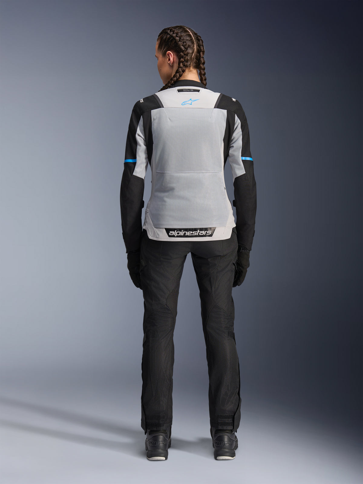 Damska kurtka Alpinestars Stella ST-2 Air, kolor Ice Gray Black, widok z tyłu na modelce, duży jasnoszary siatkowy panel na plecach, czarne rękawy z niebieskimi akcentami, logo Alpinestars na dole pleców, czarne techniczne spodnie i buty motocyklowe, letnia odzież turystyczna