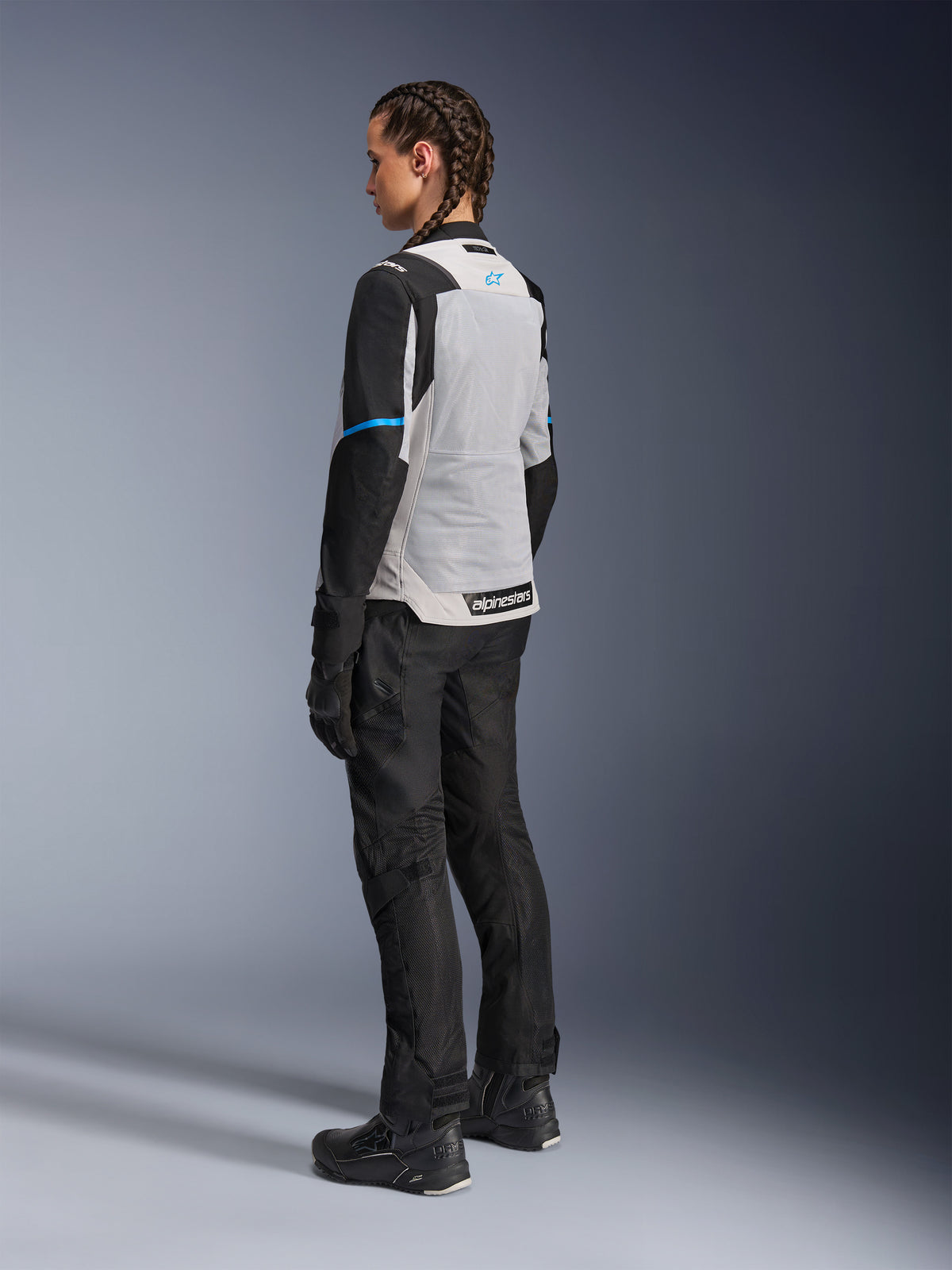Motocyklistka ubrana w kurtkę Alpinestars Stella ST-2 Air w kolorze Ice Gray i czarnym, widok z tyłu pod kątem trzech czwartych podkreślający duży jasnoszary panel siatkowy na plecach, czarne rękawy z niebieskim paskiem i niebieskim logo Alpinestars, zestawiona z czarnymi spodniami motocyklowymi, rękawicami i butami, tworząca kompletny strój do turystyki przygodowej