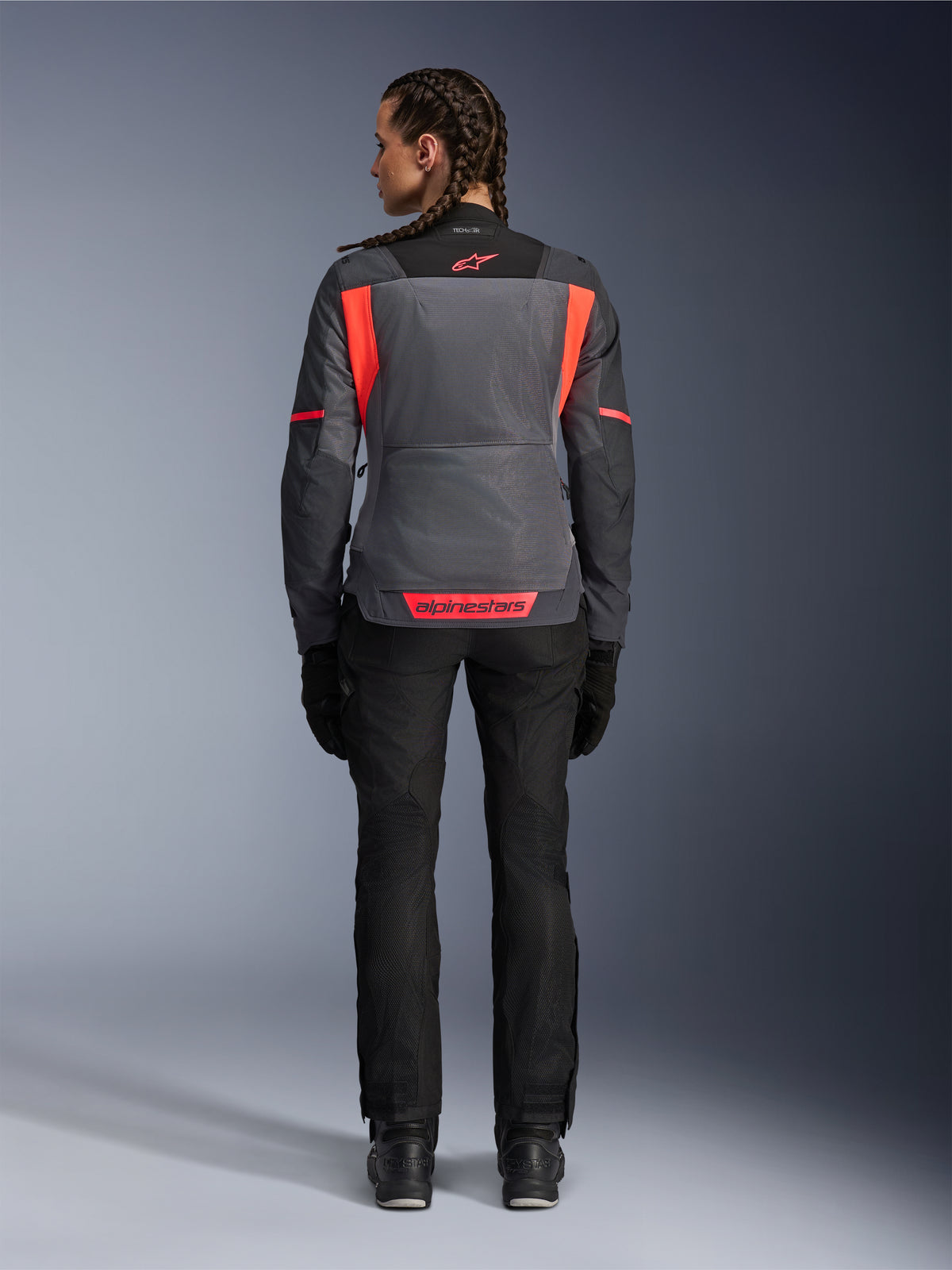 Widok od tyłu modelki ubranej w damską kurtkę Alpinestars Stella ST-2 Air w kolorach Deep Gray i Dark Gray z akcentami Fluo Coral, z dużymi siatkowymi panelami wentylacyjnymi na plecach i odblaskowymi detalami logo, w połączeniu z czarnymi spodniami motocyklowymi i rękawicami.