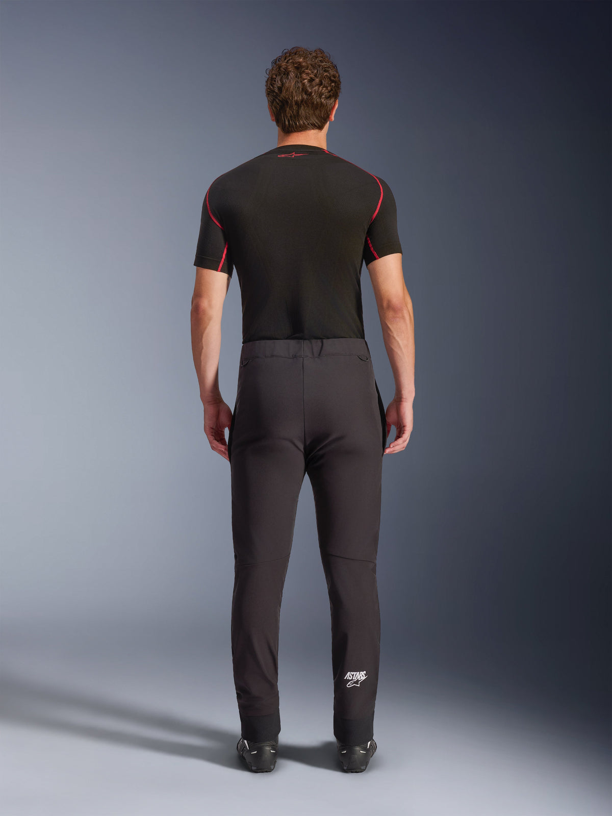 Spodnie Alpinestars Aeroshell, sportowe motocyklowe spodnie softshellowe typu crossover, czarne, widok z tyłu, zestawione z czarną warstwą bazową z krótkim rękawem, o dopasowanym kroju, profilowane kolana i białe logo Astars na dole lewej nogawki.
