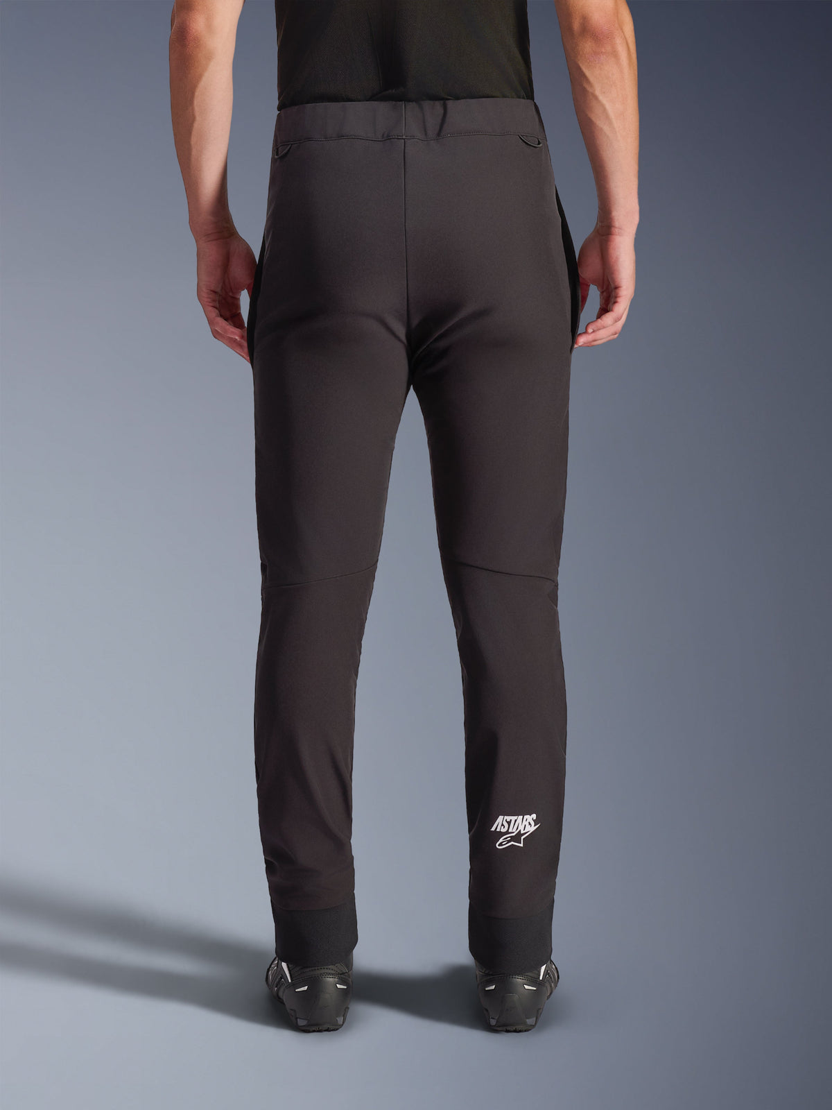Spodnie Alpinestars Aeroshell, czarne motocyklowe spodnie softshellowe, widok z tyłu prezentujący dopasowany krój i profilowane kolana, białe logo Astars na dole prawej łydki, elastyczny dolny brzeg, fason crossover do jazdy szosowej