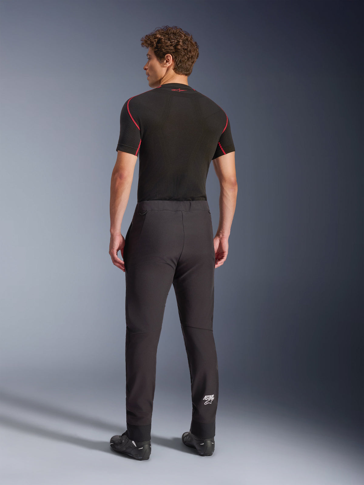 Spodnie Alpinestars Aeroshell, czarne, motocyklowe spodnie softshellowe, widok z tyłu na modelu, o nowoczesnym, zwężanym kroju, białe logo Astars na dole nogawki, elastyczne ściągacze, noszone z czarną warstwą bazową z krótkim rękawem i butami motocyklowymi.