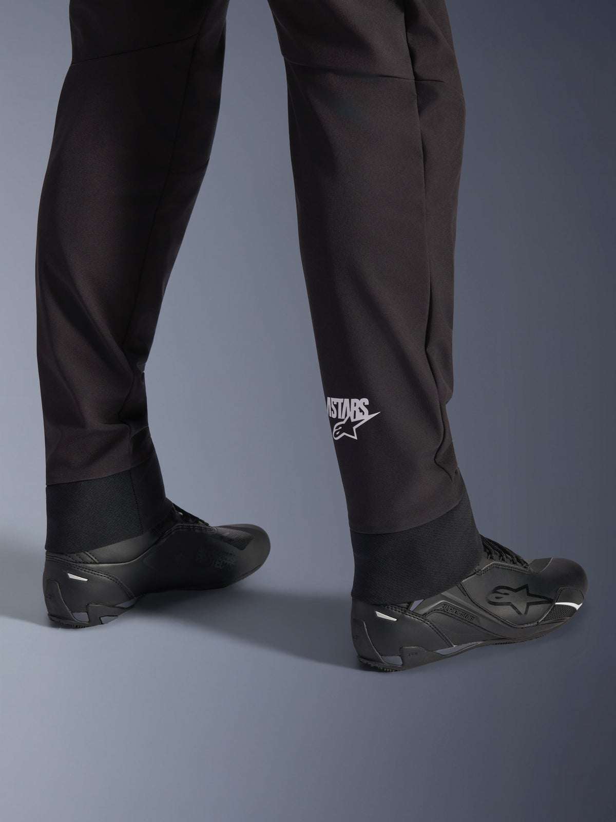 Spodnie Alpinestars Aeroshell, czarne motocyklowe spodnie softshellowe, zbliżenie na dolną część nogawki i elastyczny ściągacz, białe logo Alpinestars na łydce, zestawione z czarnymi butami motocyklowymi, nowoczesny sportowy design typu crossover