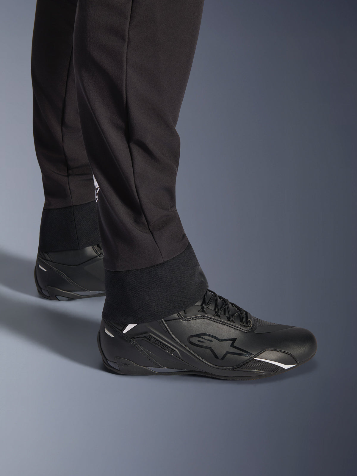Spodnie Alpinestars Aeroshell, czarne, zbliżenie na elastyczny dolny brzeg i mankiet nogawki, nowoczesny sportowy design softshell, noszone z czarnymi butami motocyklowymi Alpinestars
