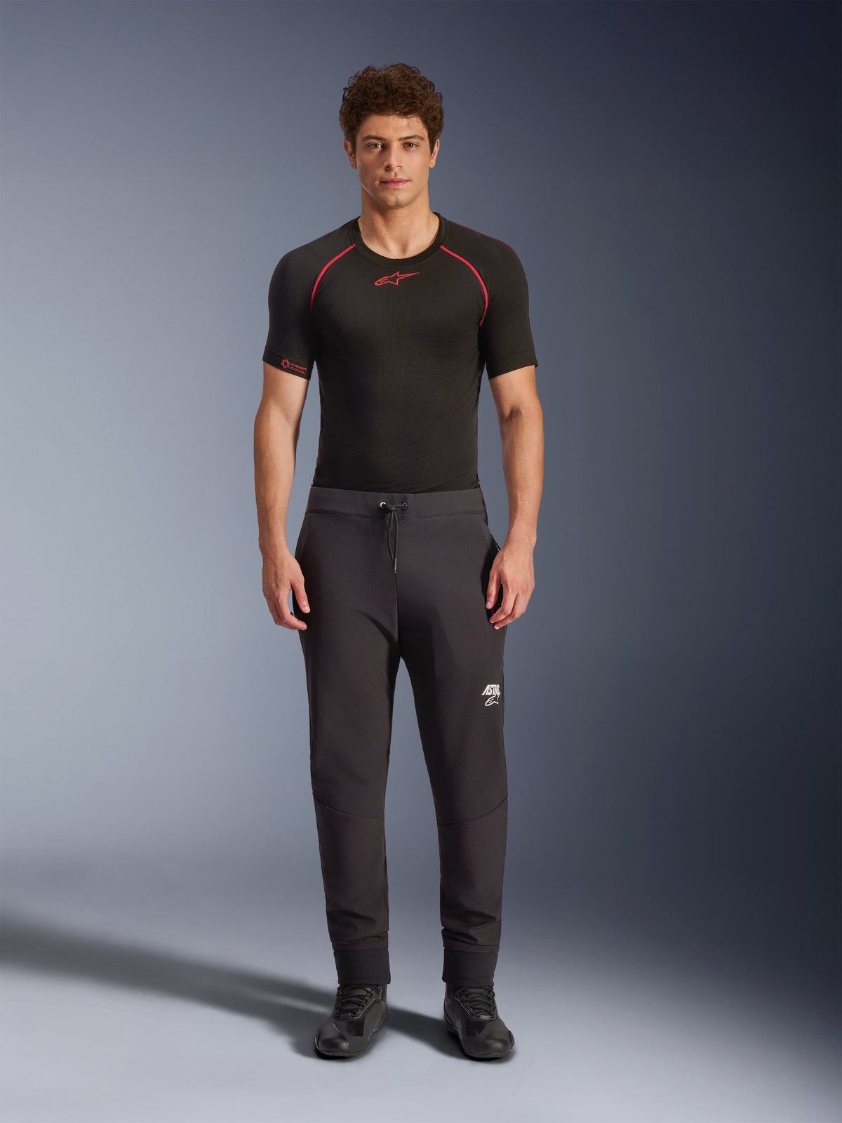 Alpinestars Aeroshell Pants, czarne motocyklowe spodnie softshellowe, noszone z czarną warstwą bazową z krótkim rękawem i czarnymi butami, elastyczny pas ze sznurkiem, białe logo Astars na udzie, elastyczne ściągacze, nowoczesna sportowa sylwetka do jazdy szosowej