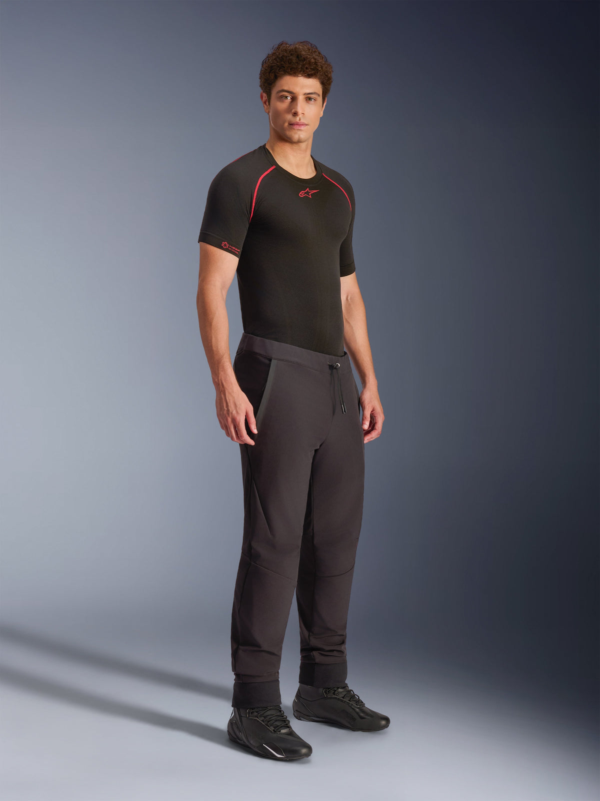 Alpinestars Aeroshell Pants, czarne, sportowe spodnie motocyklowe softshell typu crossover, prezentowane na modelu z czarną odzieżą termoaktywną i czarnymi butami, ze sznurkiem w pasie i elastycznymi ściągaczami.