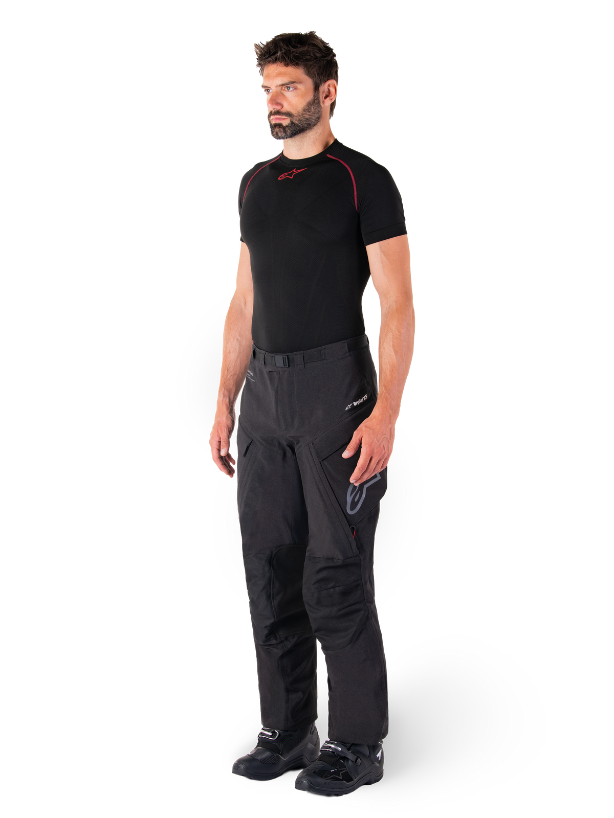 Alpinestars Hyde XT Drystar® Pants, czarne motocyklowe spodnie adventure touring prezentowane przez modela w czarnej koszulce kompresyjnej i czarnych butach, charakteryzujące się techniczną, wodoodporną konstrukcją i brandingiem Drystar®XF