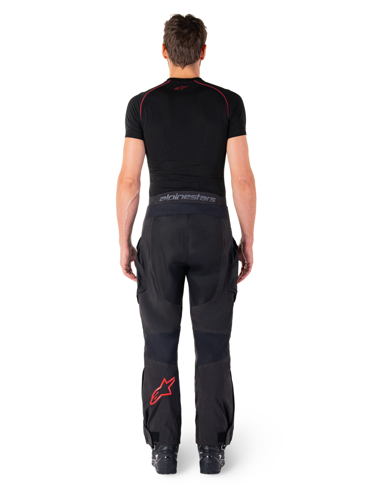 Spodnie Alpinestars Hyde Xt Drystar®, motocyklowe spodnie adventure touring, czarno-czerwone, widok z tyłu na motocyklistę w czarnej warstwie bazowej, czerwone logo gwiazdy na lewej nogawce i szary napis na pasie