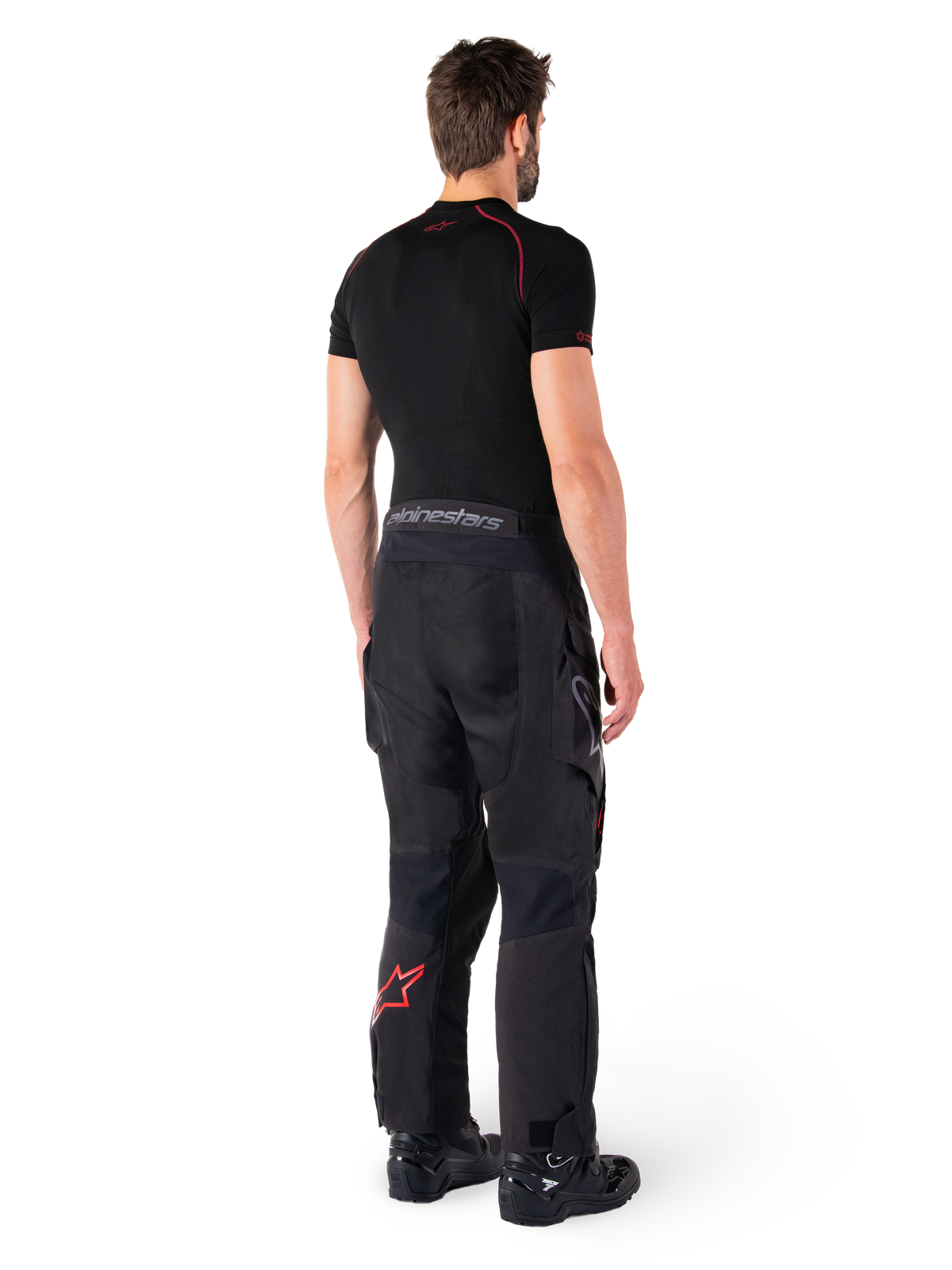 Alpinestars Hyde XT Drystar® Pants, spodnie adventure touring, czarne i jaskrawoczerwone, widok z tyłu motocyklisty ubranego w spodnie, czarną warstwę bazową i czarne buty, wytrzymała tekstylna konstrukcja, czerwone logo gwiazdy na dole nogawki, szary napis Alpinestars na pasie
