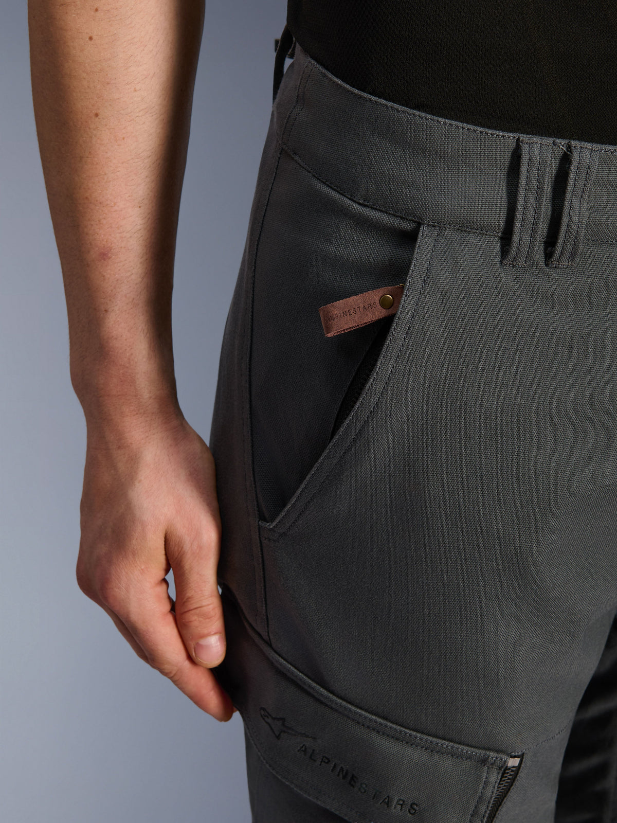 Alpinestars Borrego Drystar® Pants, motocyklowe spodnie adventure, antracytowe, zbliżenie na detal kieszeni i biodra, tekstura woskowanego płótna, brązowa skórzana metka z logo i haftowane oznaczenie Alpinestars na kieszeni cargo na udzie.