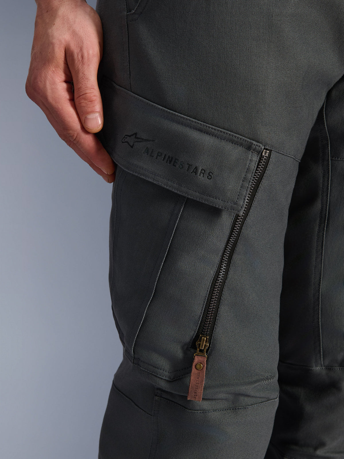 Spodnie Alpinestars Borrego Drystar®, antracytowe, zbliżenie na kieszeń cargo na udzie z haftowanym logo, teksturowany materiał z woskowanego płótna, pionowy zamek wentylacyjny z brązowym skórzanym uchwytem