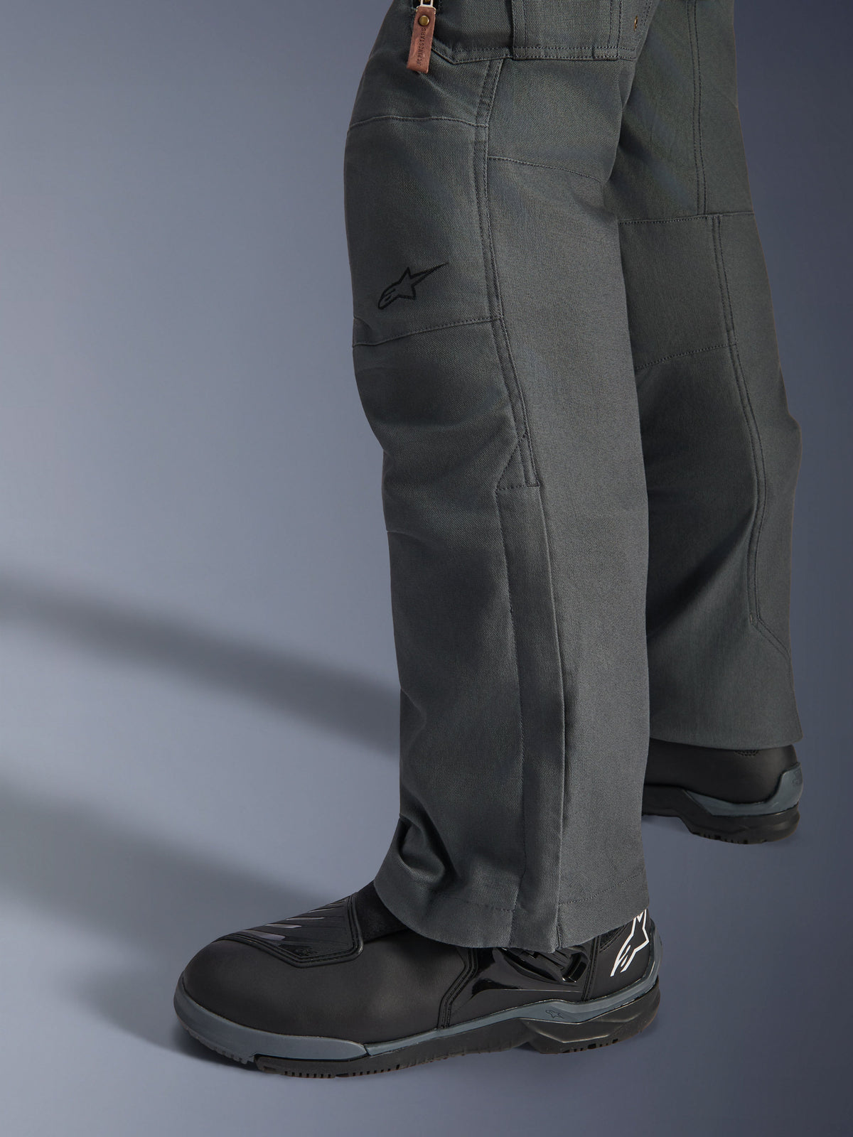Spodnie Alpinestars Borrego Drystar®, antracytowe motocyklowe spodnie adventure o wyglądzie woskowanego płótna, tekstylna konstrukcja pokazana z boku z czarnymi butami motocyklowymi, wytrzymały design do turystyki szosowej i adventure