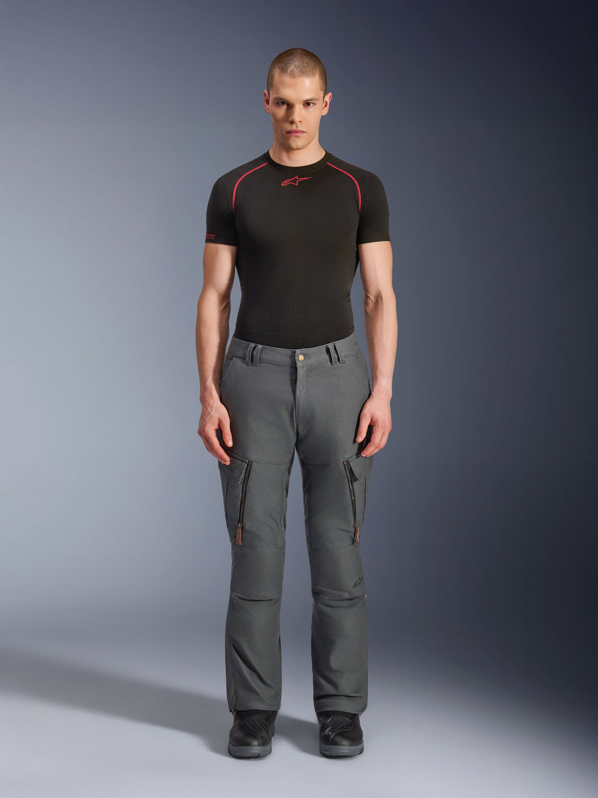 Spodnie Alpinestars Borrego Drystar®, szare spodnie motocyklowe adventure, noszone przez modela z czarno-czerwonym t-shirtem technicznym i czarnymi butami, kieszenie cargo z pionowymi zamkami, wygląd wytrzymałego woskowanego płótna, trwała konstrukcja do turystyki przygodowej i jazdy szosowej