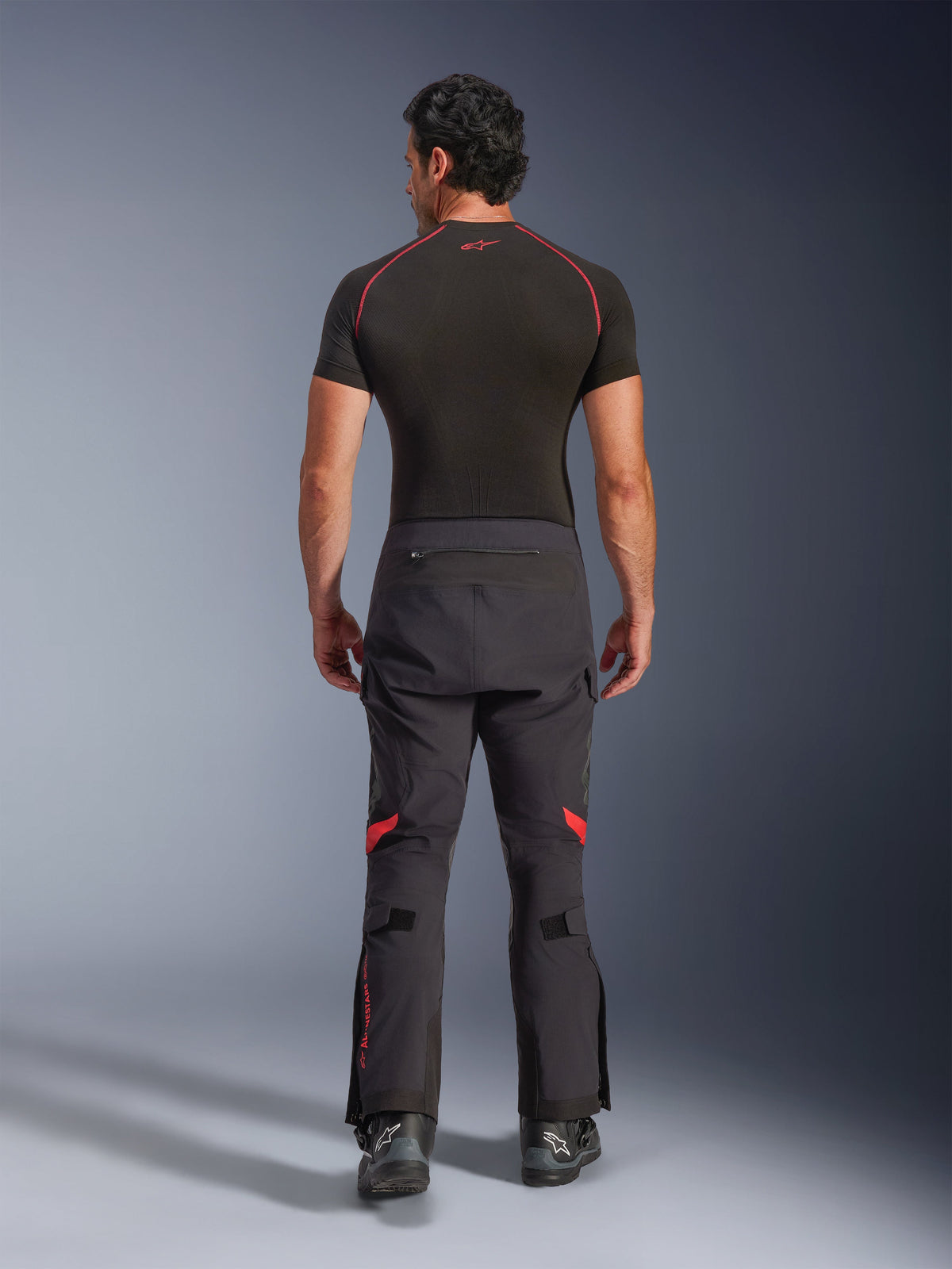 Alpinestars Halo Pro Drystar® XF Laminated Pants, motocyklowe spodnie adventure, kolor Black Dark Shadow z czerwonymi akcentami, widok z tyłu, noszone z czarną warstwą bazową z krótkim rękawem i czarnymi butami adventure, wytrzymała 3-warstwowa laminowana konstrukcja i ergonomiczne dopasowanie.