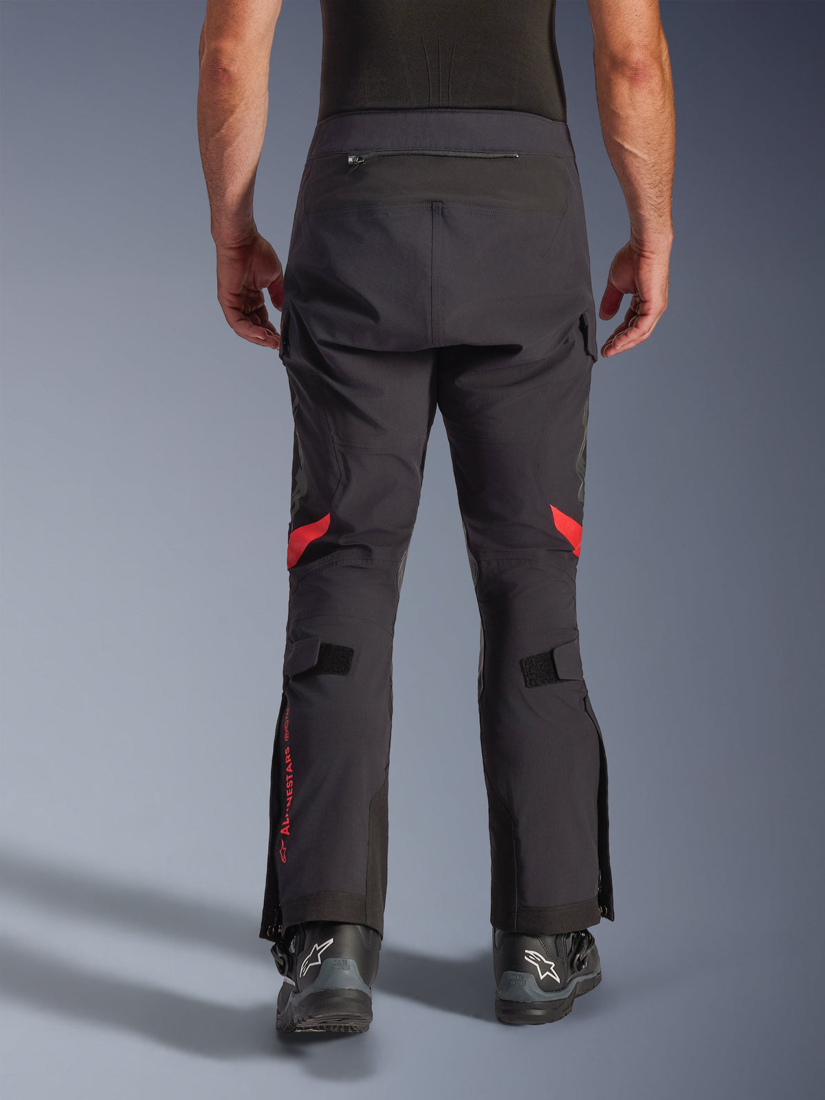 Alpinestars Halo Pro Drystar® XF Laminated Pants, Black Dark Shadow, widok od tyłu przygodowych spodni motocyklowych z czerwonymi akcentami pod kolanami i czerwonym logo na łydce, zestawione z czarnymi butami Alpinestars, wytrzymały 3-warstwowy laminowany materiał tekstylny zapewniający wydajność w każdym terenie