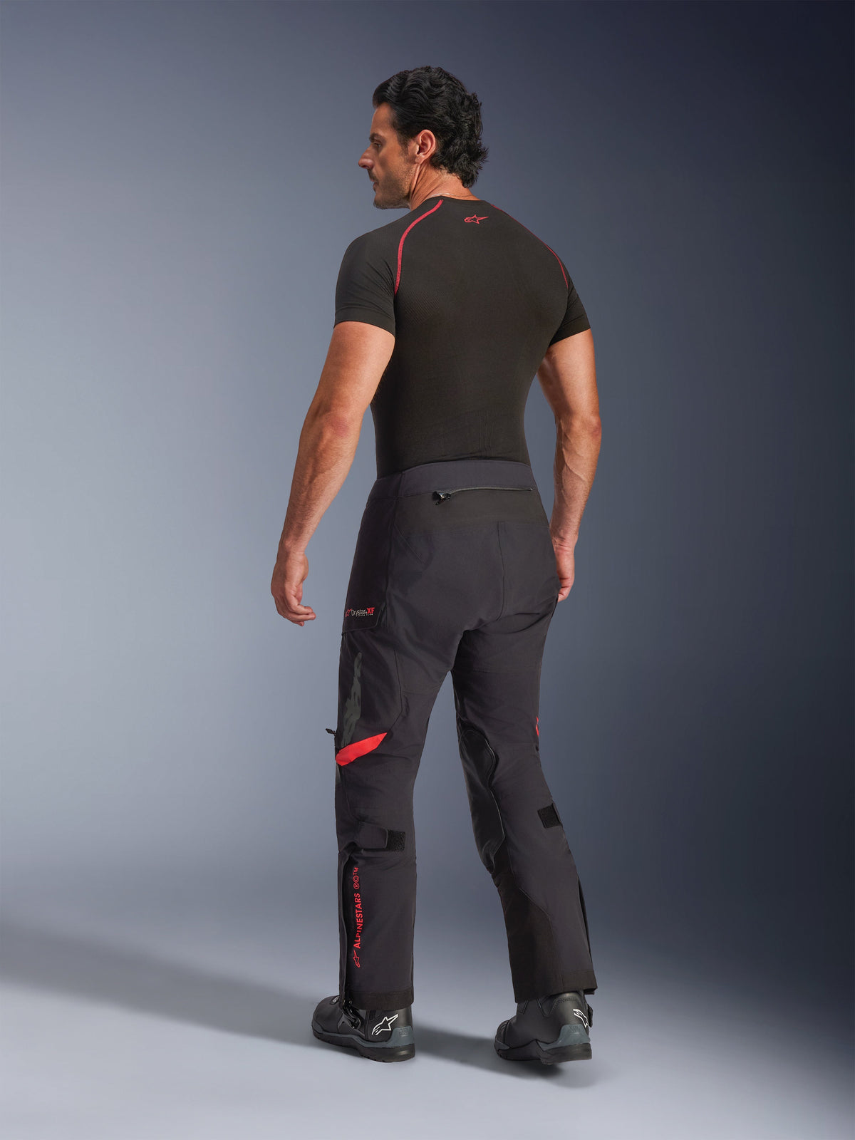 Spodnie laminowane Alpinestars Halo Pro Drystar® XF, kolor Black Dark Shadow z czerwonymi akcentami, widok z tyłu na modelu, zestawione z czarną warstwą bazową i butami motocyklowymi, techniczna odzież adventure touring do jazdy w każdym terenie