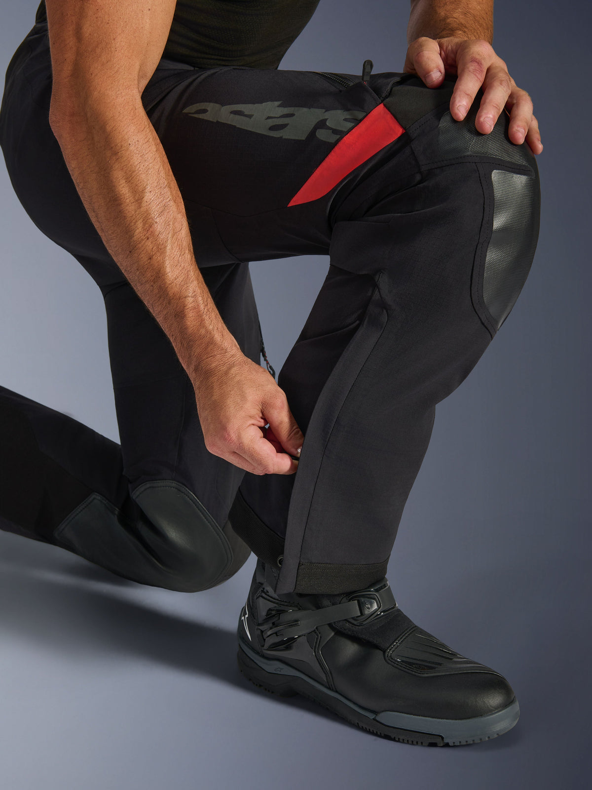Alpinestars Halo Pro Drystar® XF Laminated Pants, Black Dark Shadow, zbliżenie na dolną część nogawki z regulowanym zamkiem na łydce, z trwałymi wzmocnieniami na kolanach, czerwonymi akcentami i brandingiem „astars”, noszone z czarnymi butami motocyklowymi