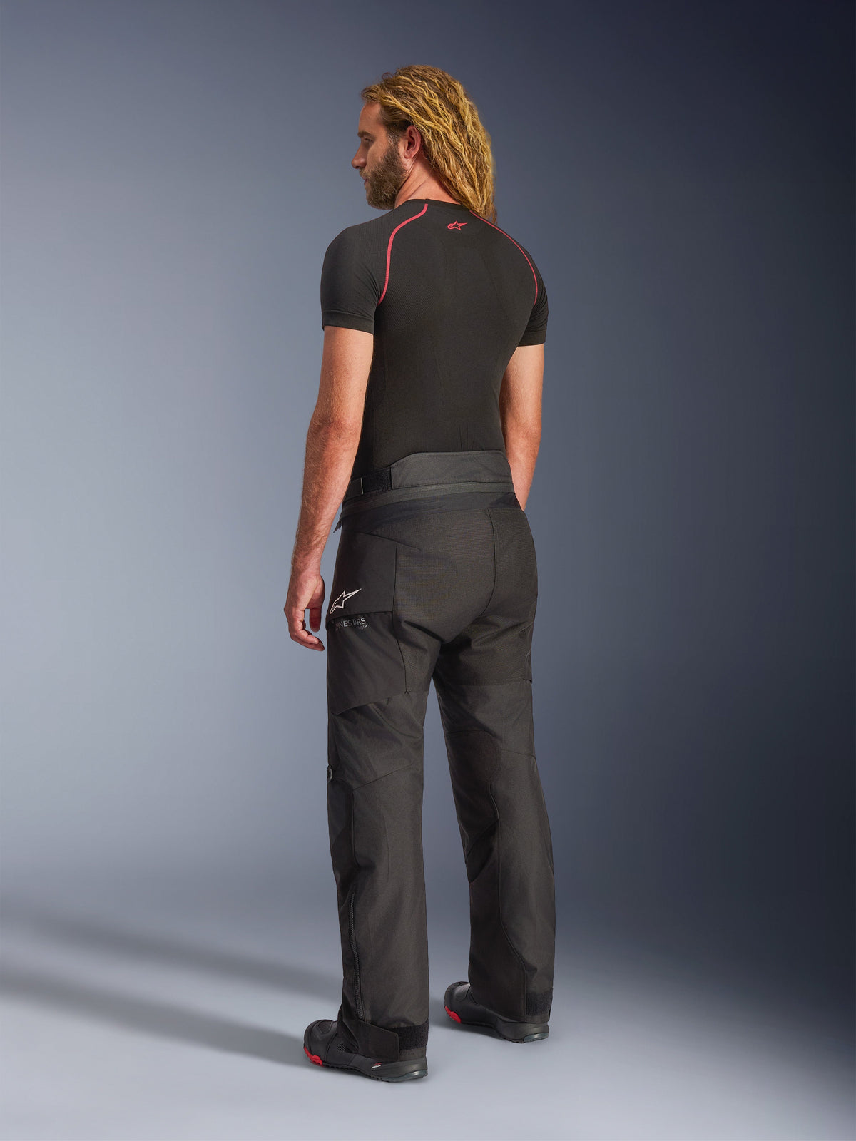 Alpinestars Andes V4 Drystar® Pants, Black motorcycle adventure touring pants, worn by a model from a rear-side view, featuring a white Alpinestars logo on the hip, paired with a black short-sleeve base layer and riding boots, durable textile construction for road and adventure use.