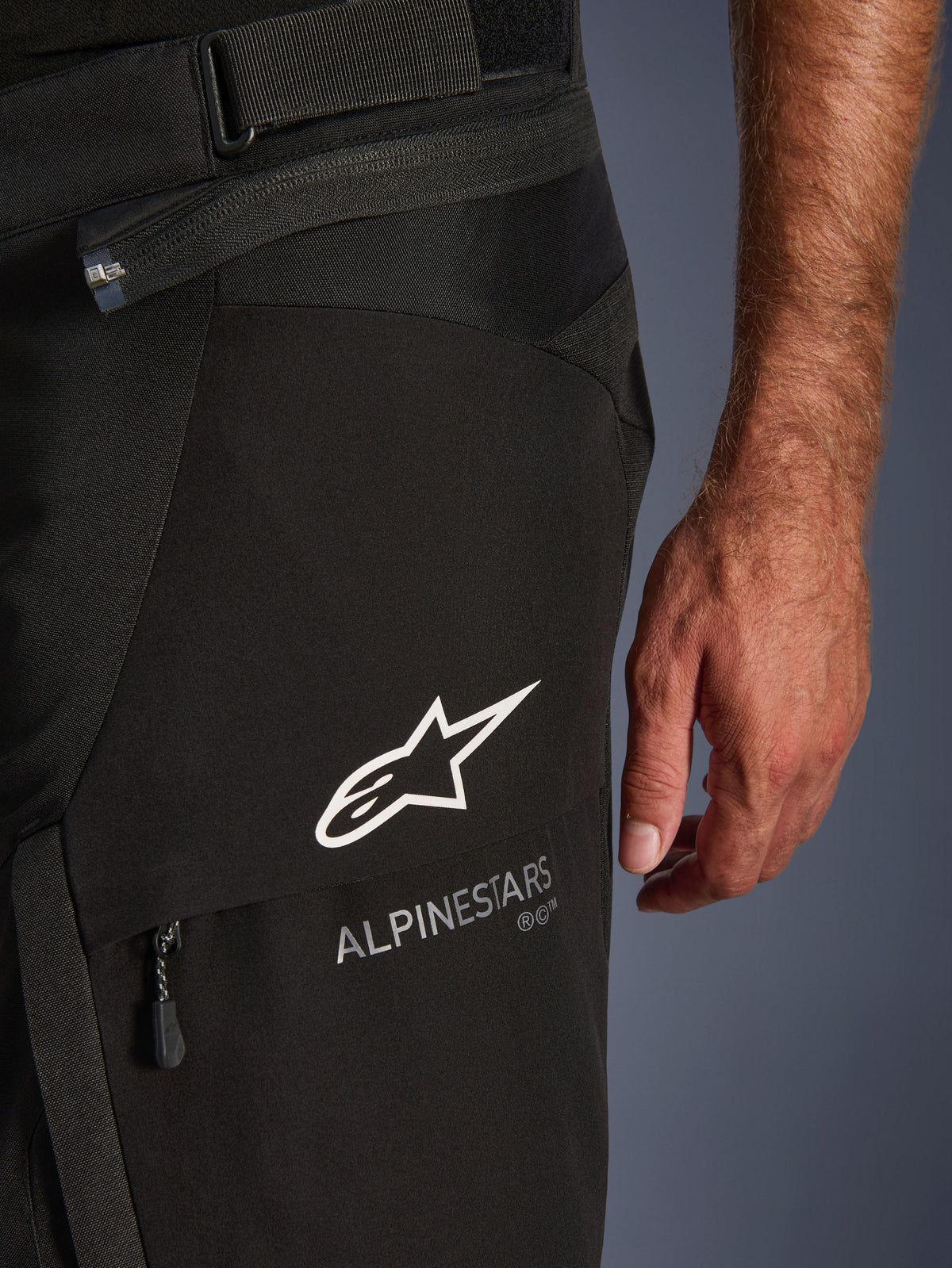Close-up detail of Alpinestars Andes V4 Drystar® Pants in Black, featuring the white Alpinestars logo and wordmark on a cargo pocket flap, with visible waist adjustment strap and connection zipper, designed for adventure motorcycle touring.