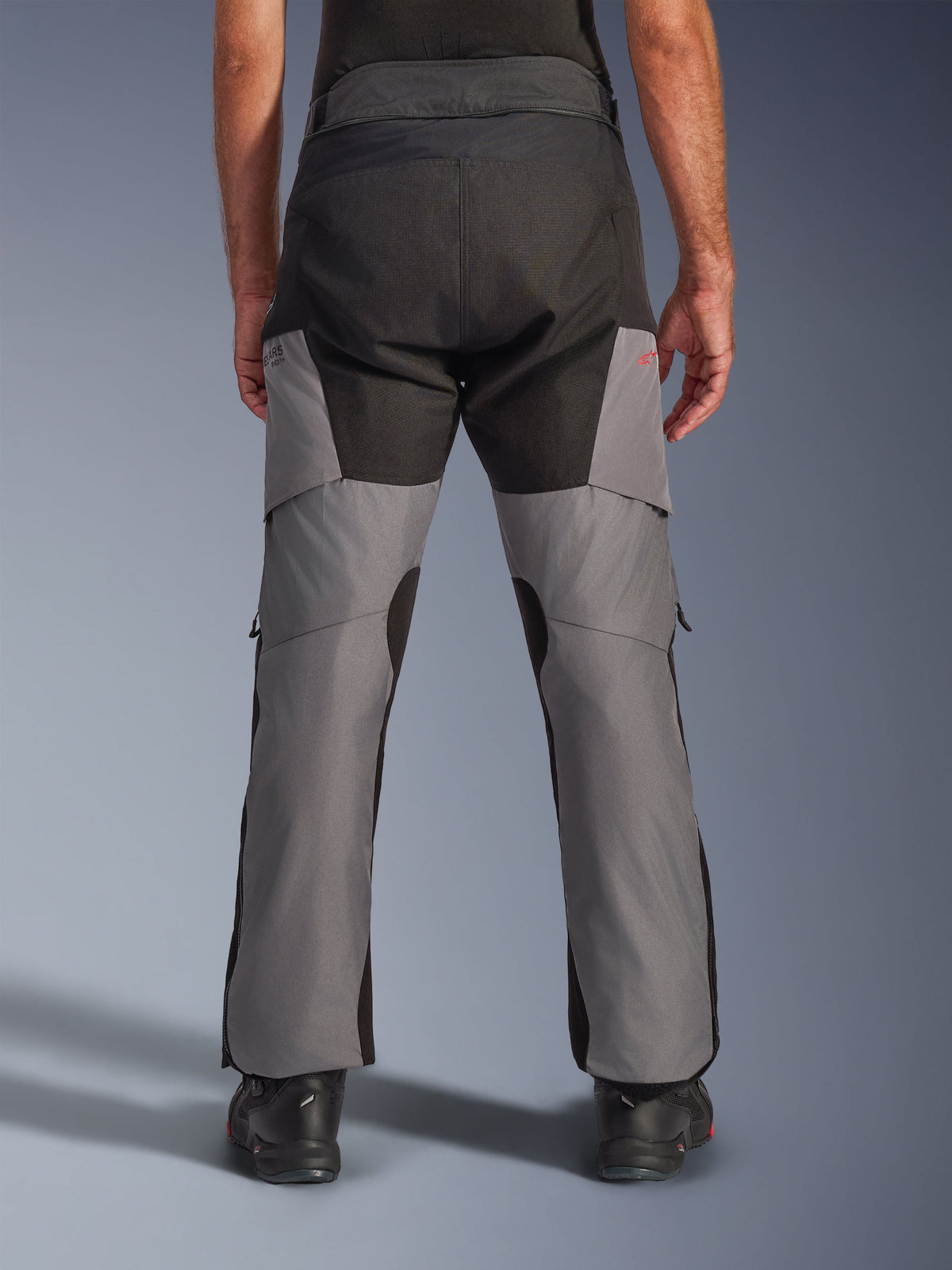 Alpinestars Andes V4 Drystar® Pants, przygodowe spodnie motocyklowe, czarno-ciemnoszare, widok z tyłu prezentujący wzmocnione czarne siedzenie i szare nogawki, małe czerwone logo, połączone z czarnymi butami motocyklowymi, wytrzymała tekstylna konstrukcja do turystyki w każdą pogodę