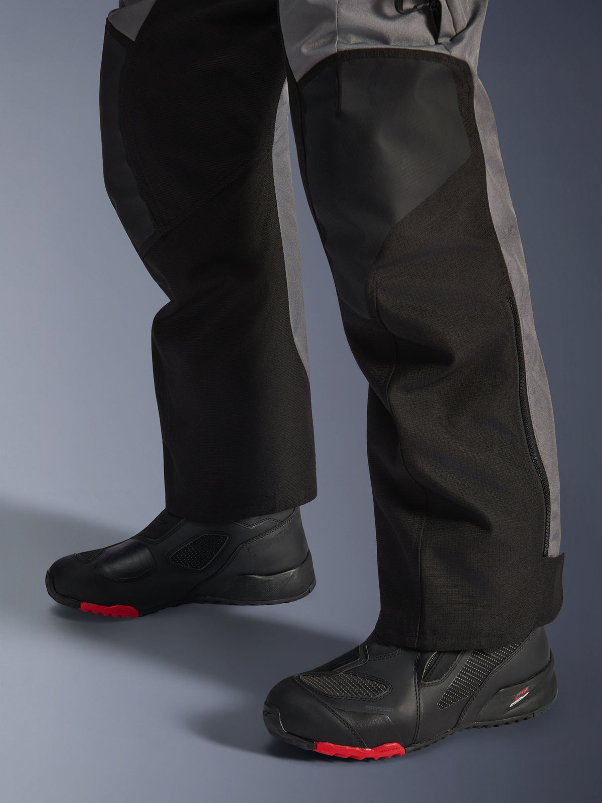 Alpinestars Andes V4 Drystar® Pants, tekstylne spodnie motocyklowe, czarne i ciemnoszare, noszone z czarnymi butami motocyklowymi z czerwonymi podeszwami, design adventure touring ze wzmocnionymi panelami na nogawkach i prostym krojem.