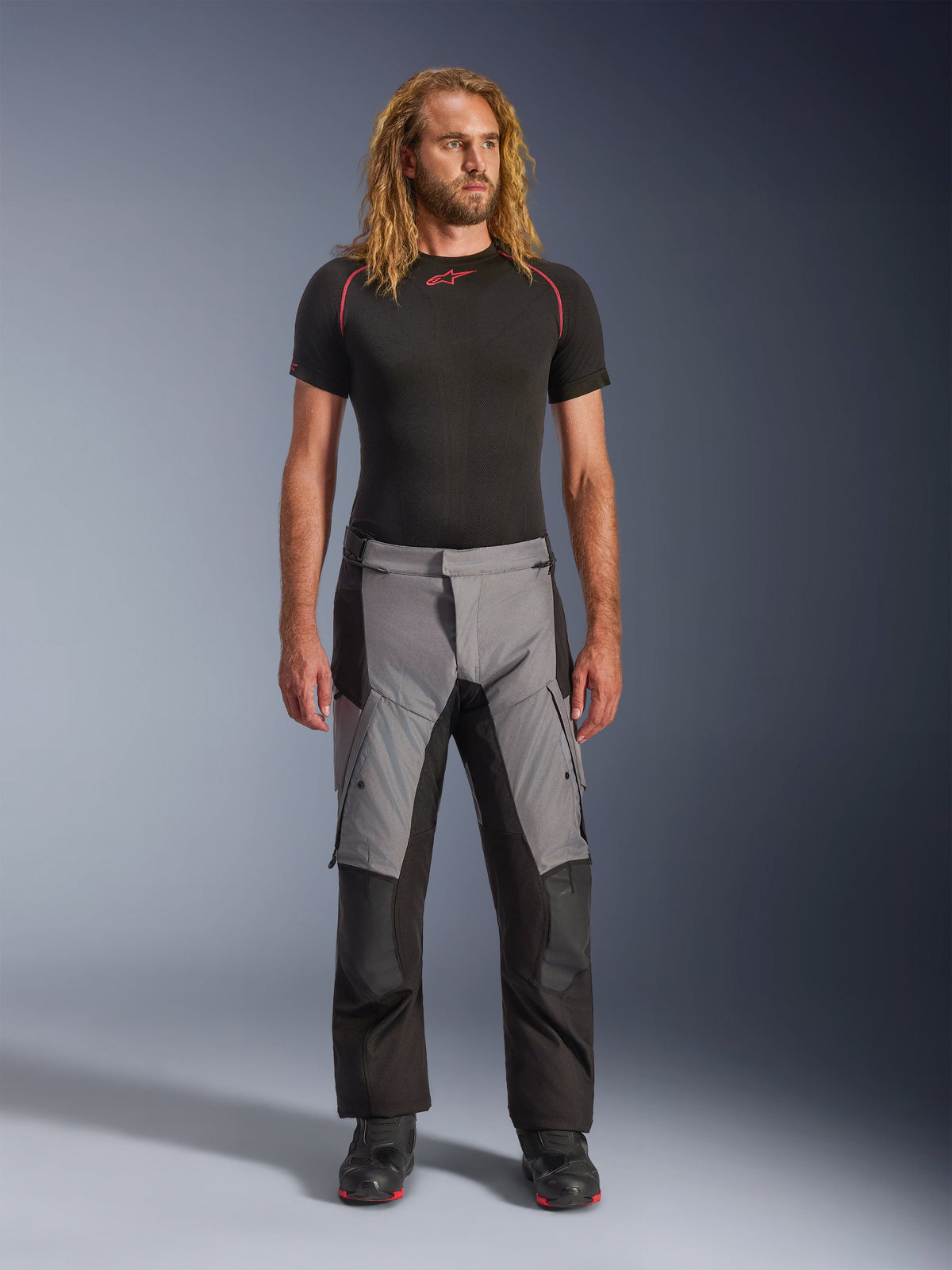 Alpinestars Andes V4 Drystar® Pants, czarno-ciemnoszare, spodnie motocyklowe adventure prezentowane przez modela w czarnej bieliźnie termoaktywnej i czarnych butach, wielopanelowa konstrukcja tekstylna z szarymi przednimi sekcjami i czarnymi wzmocnieniami, przeznaczone do turystyki i jazdy szosowej w każdą pogodę