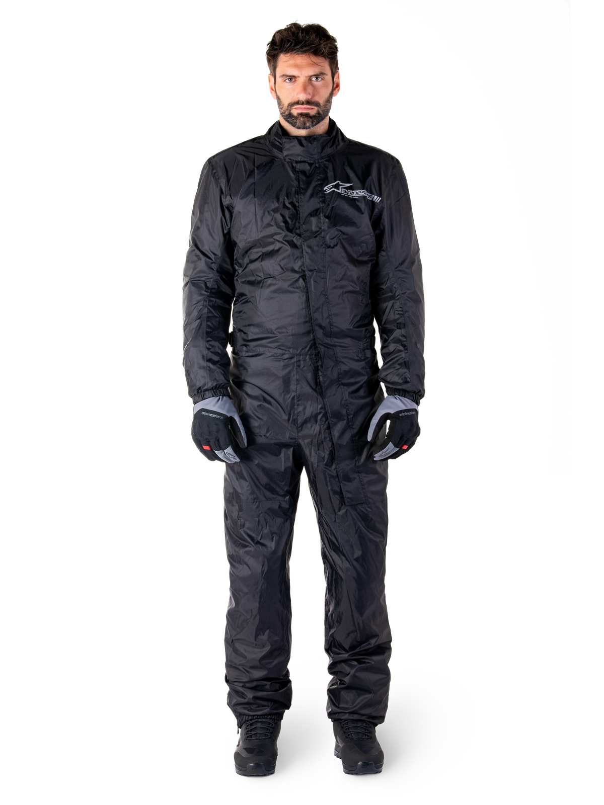 Alpinestars Hurricane Rain V2 Suit, pełny motocyklowy kombinezon przeciwdeszczowy w kolorze czarnym, wodoodporna odzież ochronna z wysokim kołnierzem i przednim zapięciem uszczelniającym, białe logo na piersi, prezentowany z czarno-szarymi rękawicami i butami