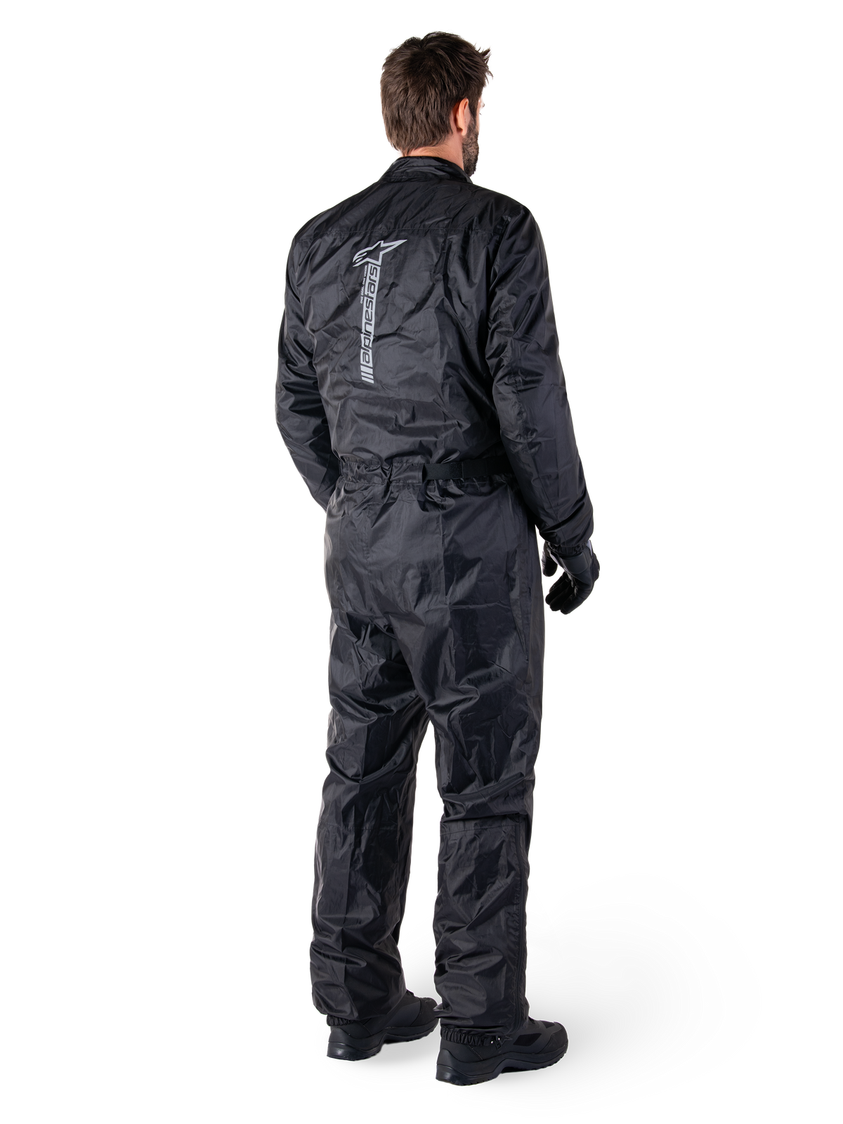 Alpinestars Hurricane Rain V2 Suit, czarny jednoczęściowy wodoodporny motocyklowy kombinezon przeciwdeszczowy, widok z tyłu z dużym pionowym odblaskowym logo, lekka wiatroodporna tkanina, noszony z czarnymi rękawicami i butami