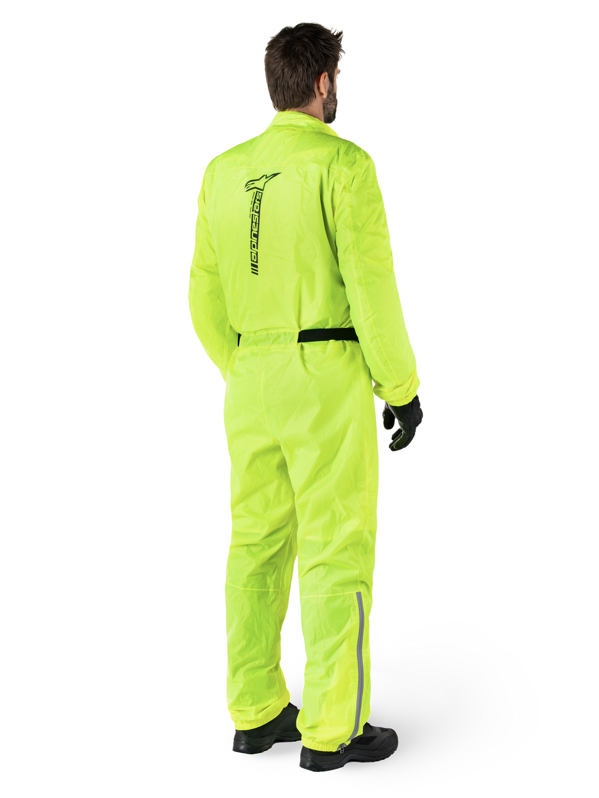 Alpinestars Hurricane Rain V2 Suit, Yellow Fluo, widok z tyłu motocyklisty w jednoczęściowym motocyklowym kombinezonie przeciwdeszczowym, z czarnym logo Alpinestars na plecach, czarnym paskiem w talii i odblaskowymi paskami na dolnej części nogawek, noszony z czarnymi rękawicami i butami