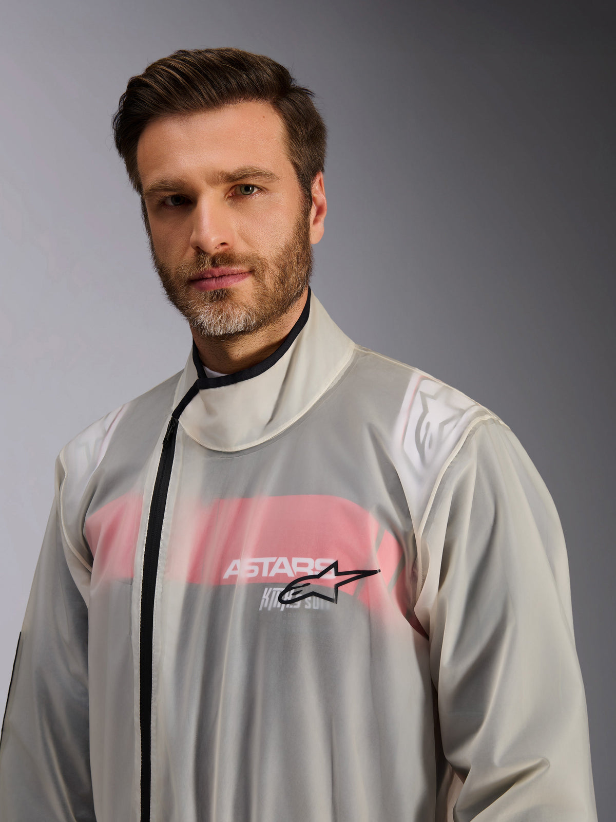 Alpinestars Kart Rain Suit, kombinezon przeciwdeszczowy do kartingu, przezroczysty, noszony przez mężczyznę, wodoodporny transparentny materiał ukazujący czerwono-biały kombinezon wyścigowy pod spodem, wysoki kołnierz z czarnym zamkiem błyskawicznym i logo Alpinestars na klatce piersiowej