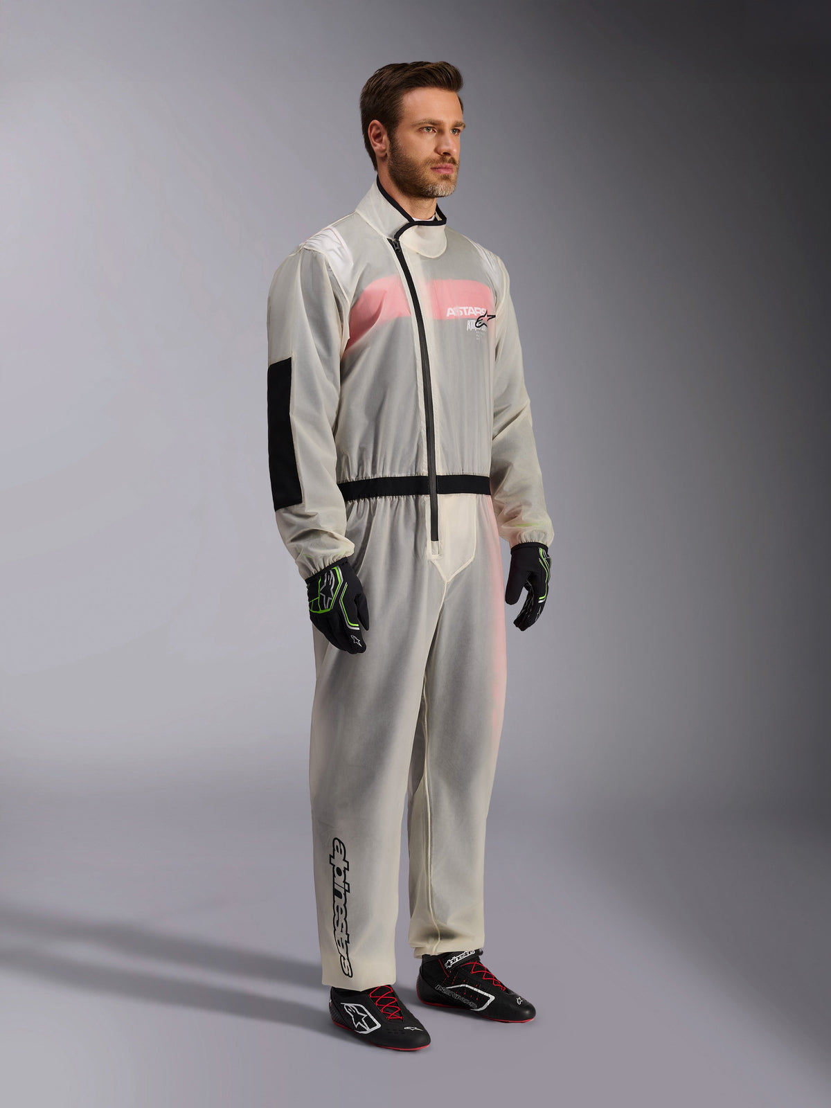 Alpinestars Kart Rain Suit, przezroczysty wodoodporny kombinezon wierzchni do kartingu, półprzezroczysty materiał ukazujący różowo-biały kombinezon wyścigowy pod spodem, z czarnym asymetrycznym zamkiem, wzmocnioną łatą na łokciu i czarnym pasem, noszony z czarnymi rękawicami kartingowymi i butami wyścigowymi.