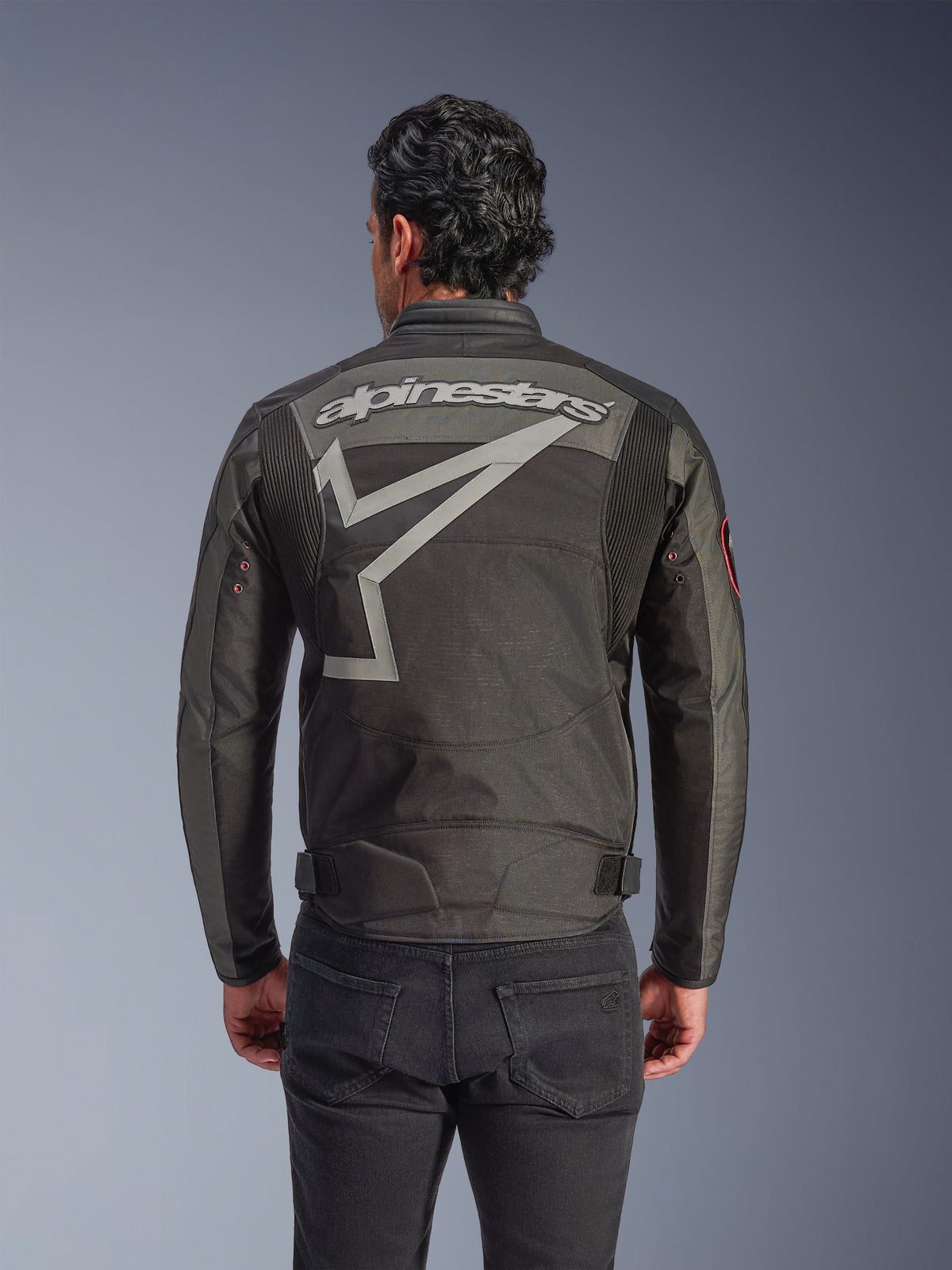 Kurtka Alpinestars Axton, męska kurtka motocyklowa, czarna, antracytowa, czerwona i mandarynkowa, widok z tyłu z dużym szarym logotypem Alpinestars i stylizowaną grafiką gwiazdy, harmonijkowe panele na ramionach oraz metalowe oczka w stylu vintage na rękawach