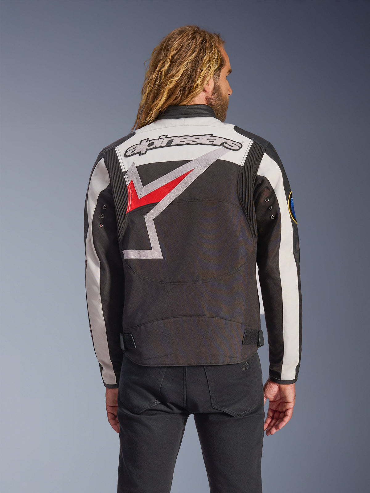 Kurtka Alpinestars Axton, kurtka motocyklowa, czarna, złamana biel i jasna czerwień, widok z tyłu z dużym, otaczającym logo gwiazdy i znakiem słownym, design inspirowany stylem vintage z akordeonowymi panelami na ramionach i regulacją w pasie.