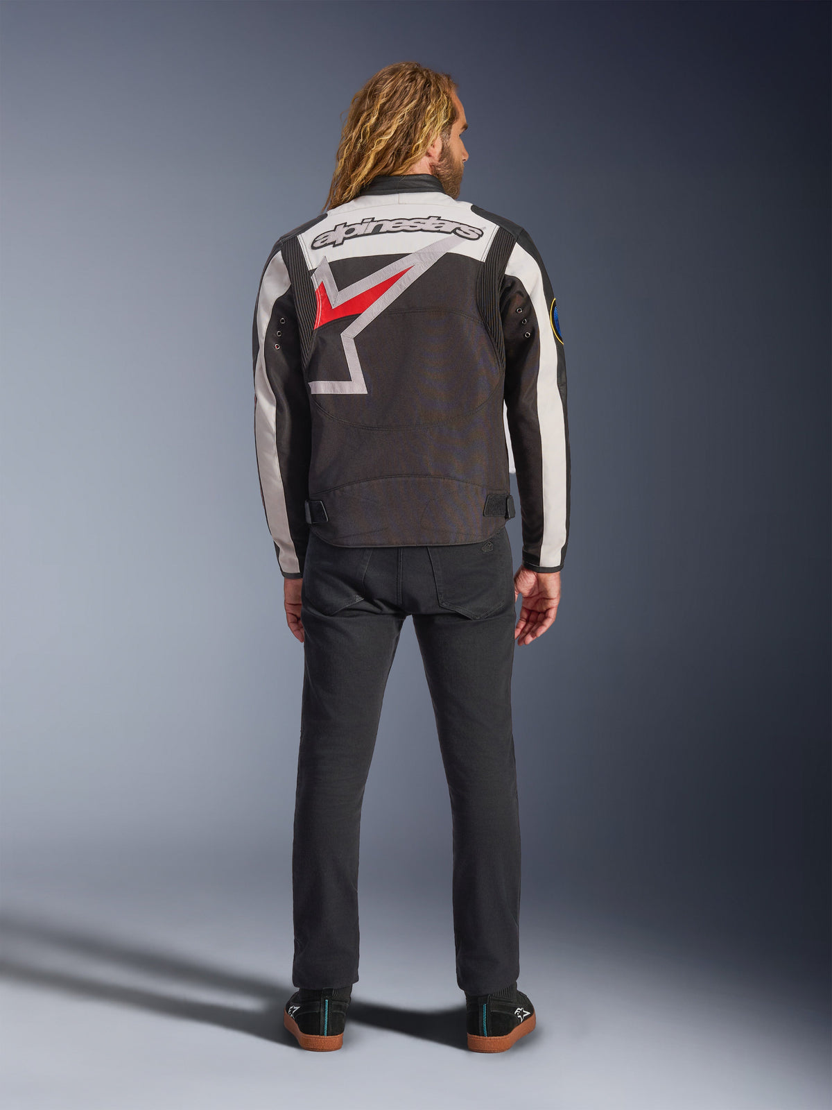 Kurtka Alpinestars Axton, kurtka motocyklowa, czarno-biała (off-white) z jaskrawoczerwonymi elementami, widok z tyłu na modelu, duże logo gwiazdy Alpinestars na plecach, kremowe panele na rękawach, czarne spodnie, design inspirowany odzieżą wyścigową w stylu vintage
