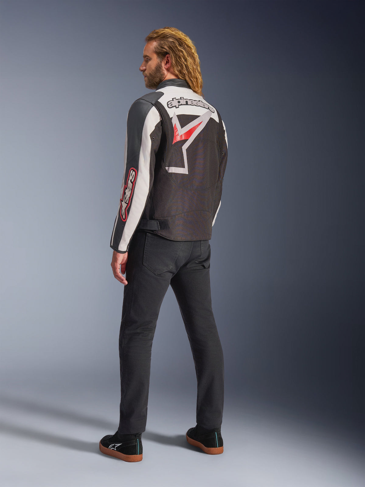 Kurtka motocyklowa Alpinestars Axton, czarna z brudnobiałymi i jaskrawoczerwonymi akcentami, widok od tyłu na modelu, duże logo gwiazdy i napis Alpinestars na plecach, design inspirowany stylem vintage z panelami akordeonowymi, czarne spodnie i buty sportowe