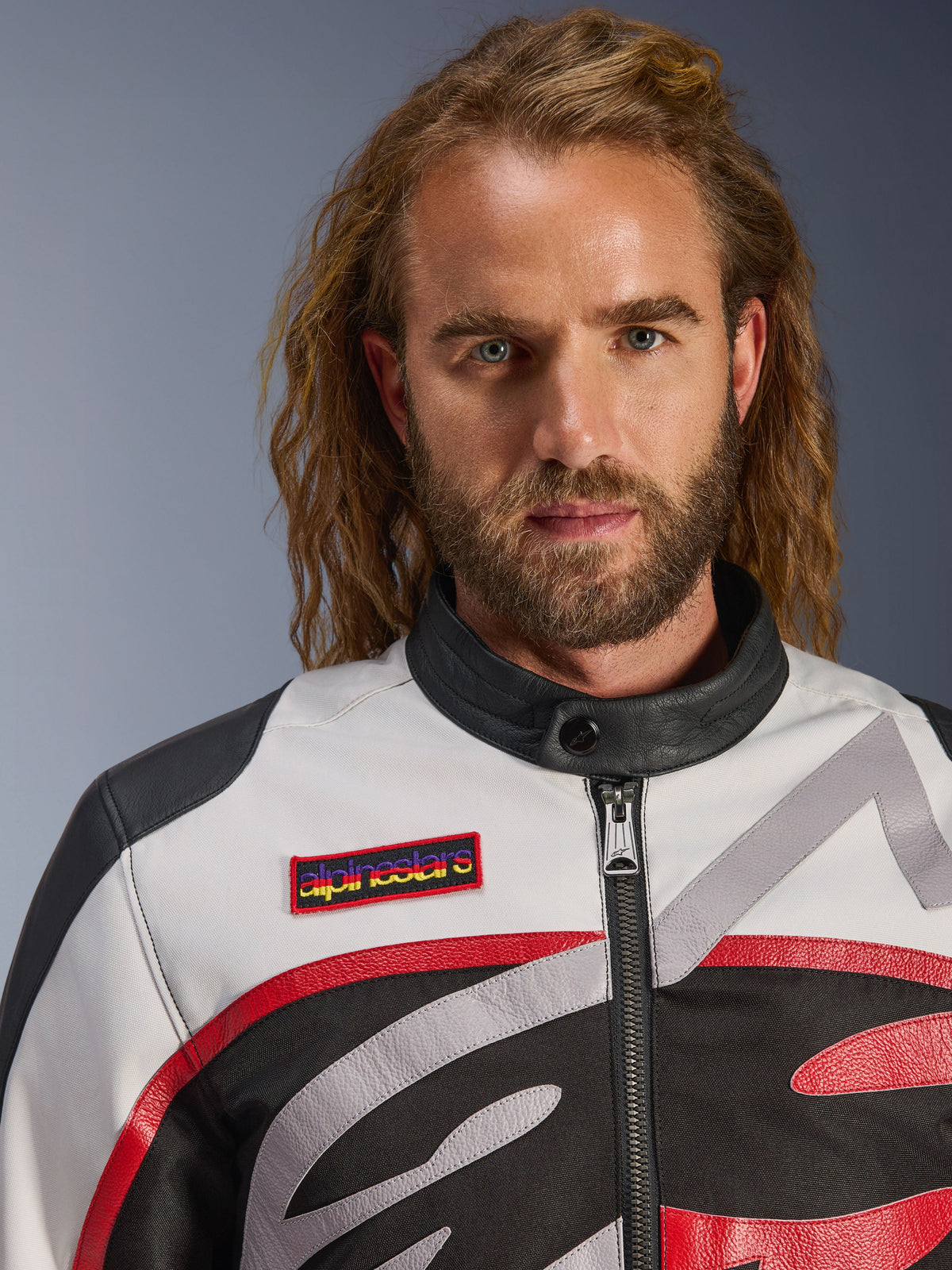Mężczyzna w kurtce Alpinestars Axton, motocyklowa kurtka inspirowana stylem vintage w kolorach czarnym, złamanej bieli i jasnej czerwieni, zbliżenie na markową naszywkę na piersi i minimalistyczny kołnierz motocyklowy, współczesna odzież do jazdy szosowej