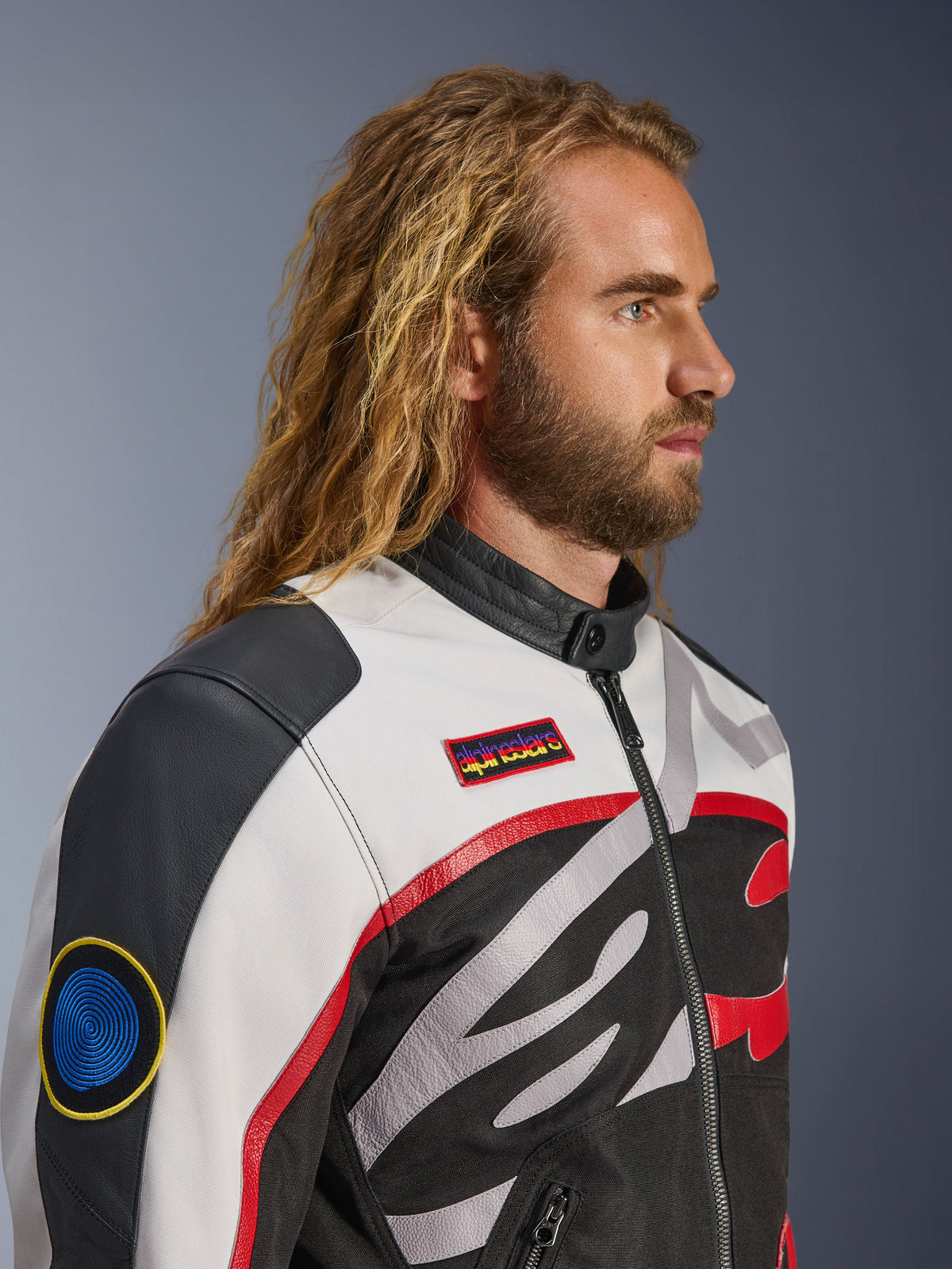 Mężczyzna z długimi włosami i brodą w kurtce Alpinestars Axton, kurtka motocyklowa, kolor czarny, złamana biel i jaskrawy czerwony, design inspirowany stylem vintage wyścigowym z markowymi naszywkami i logo oplatającym ciało, zbliżenie z profilu, współczesny styl miejski