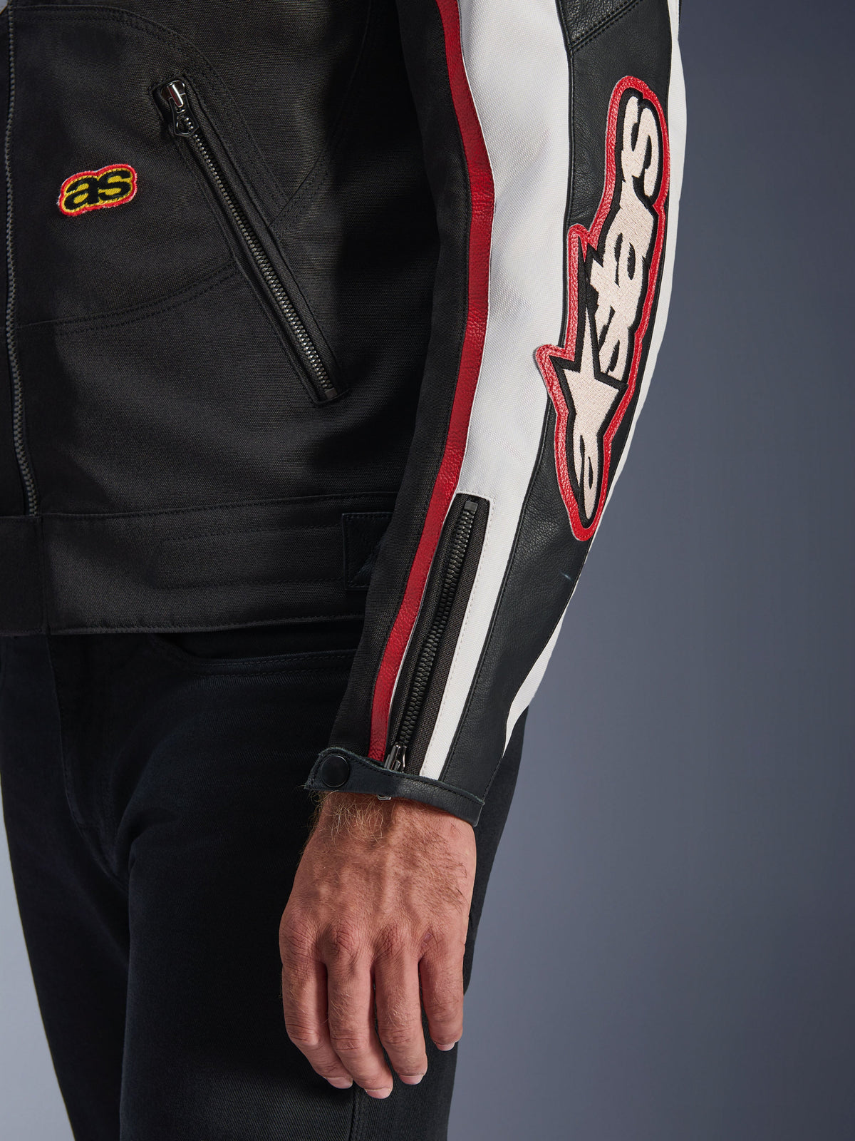 Kurtka Alpinestars Axton, zbliżenie na rękaw i tors, czarna z akcentami w kolorze złamanej bieli i jaskrawoczerwonym, duże haftowane logo Alpinestars na ramieniu, pionowe pasy wyścigowe, mankiet zapinany na zamek i zatrzask, wytrzymała konstrukcja tekstylna