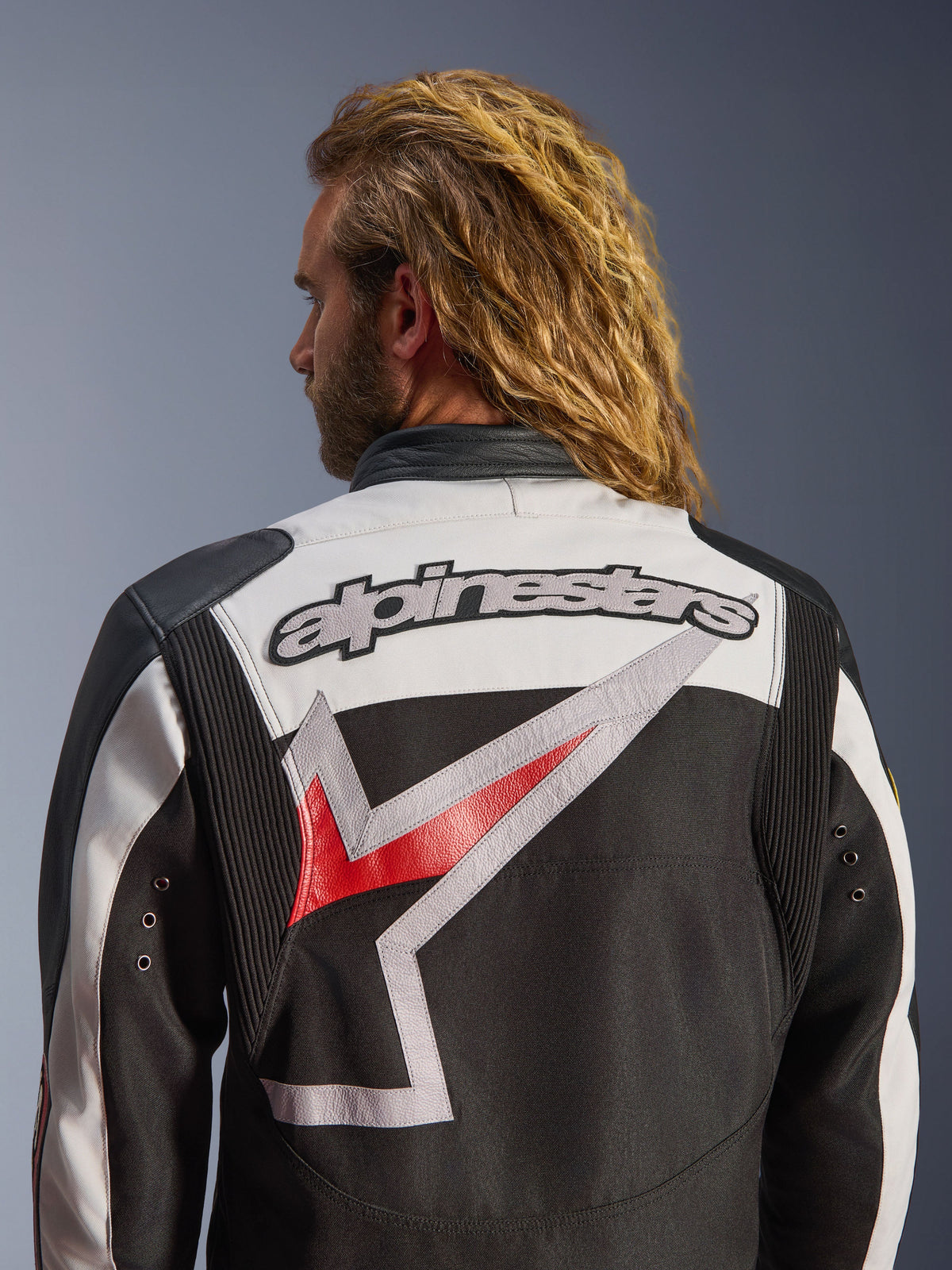Kurtka motocyklowa Alpinestars Axton, czarna, złamana biel i jaskrawa czerwień, widok z tyłu z dużym haftowanym logo Alpinestars i stylizowaną grafiką, harmonijkowe panele na ramionach dla większej swobody ruchów, design inspirowany stylem vintage z metalowymi detalami w formie oczek