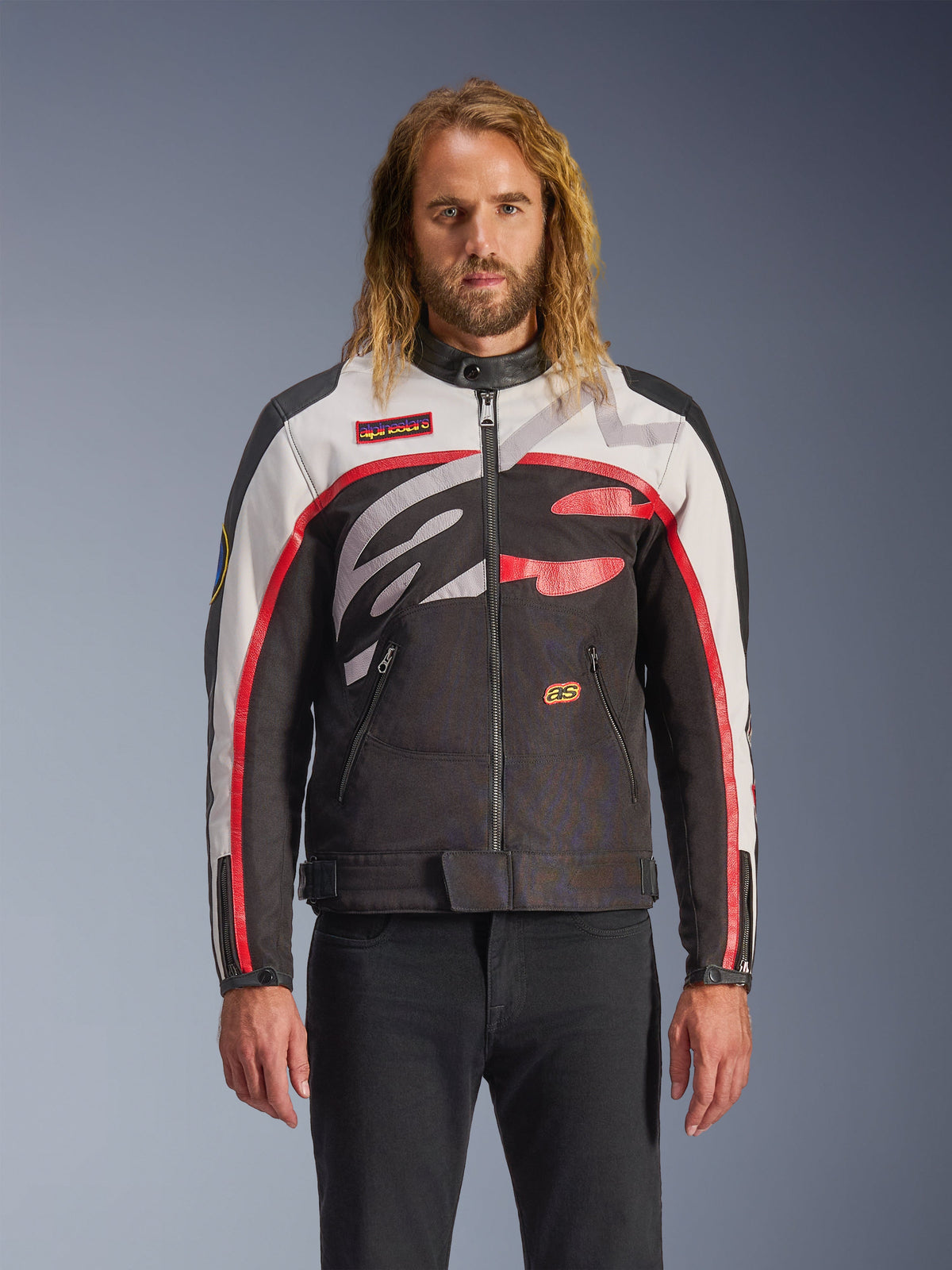 Alpinestars Axton Jacket, kurtka motocyklowa, czarna, złamana biel i jaskrawa czerwień, z wyrazistymi abstrakcyjnymi wzorami na klatce piersiowej i owijanym logo, czerwone paski na rękawach, design inspirowany stylem vintage z minimalistycznym kołnierzem, noszona przez modela z czarnymi jeansami