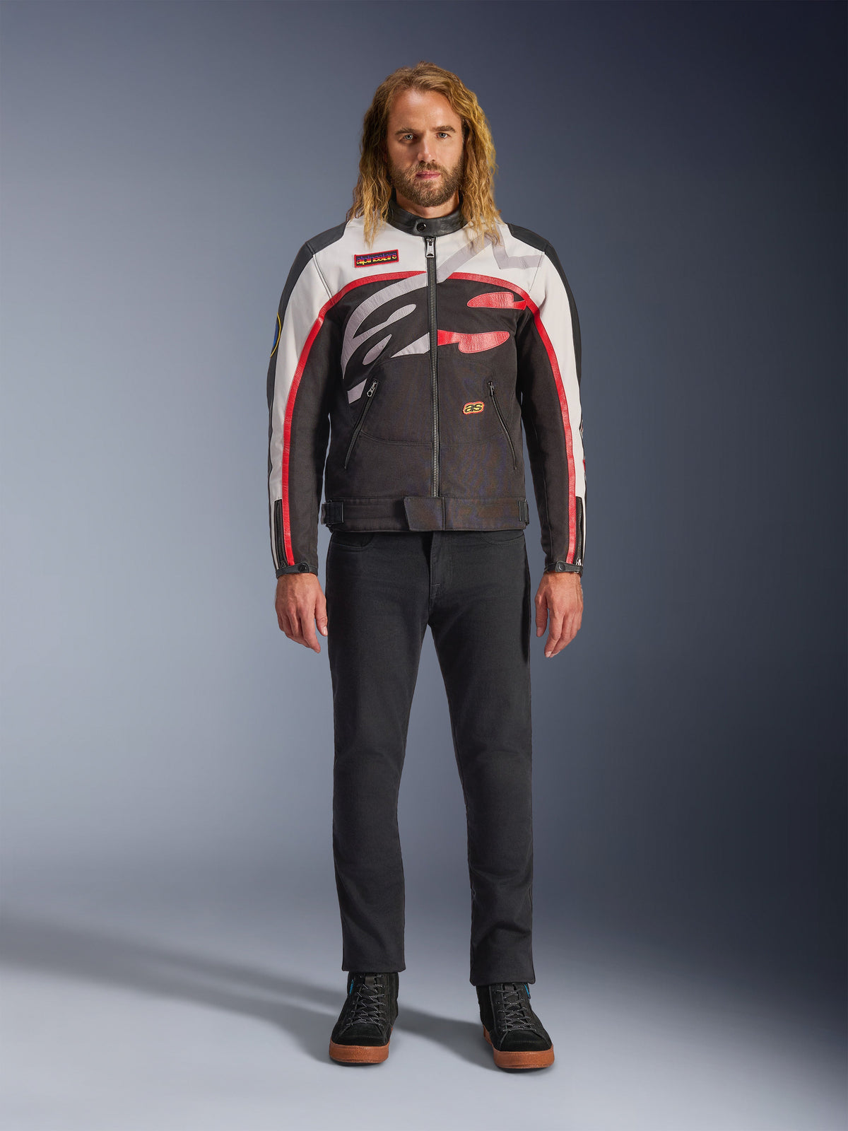 Kurtka Alpinestars Axton, kurtka motocyklowa, czarna, złamana biel i jaskrawa czerwień, prezentowana przez modela w czarnych spodniach i trampkach, z odważnym, oplatającym logo i designem inspirowanym stylem wyścigowym vintage