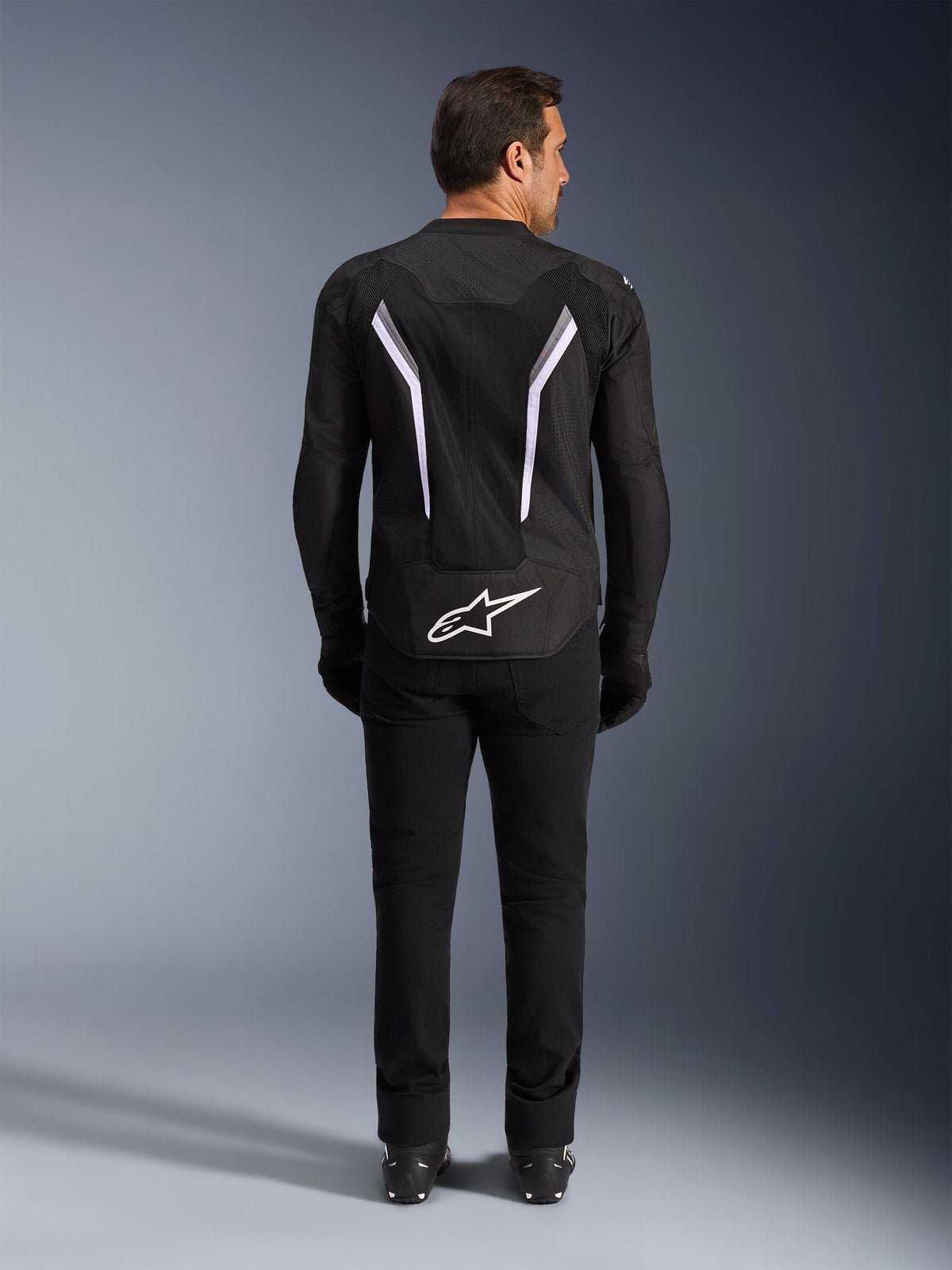 Kurtka Alpinestars AST V3 Air, widok z tyłu, czarna i ciemnoszara z białymi akcentami, duże białe logo Astar na dole, odblaskowe paski na plecach, siatkowa konstrukcja tekstylna, prezentowana z czarnymi spodniami i butami w sportowym stylu