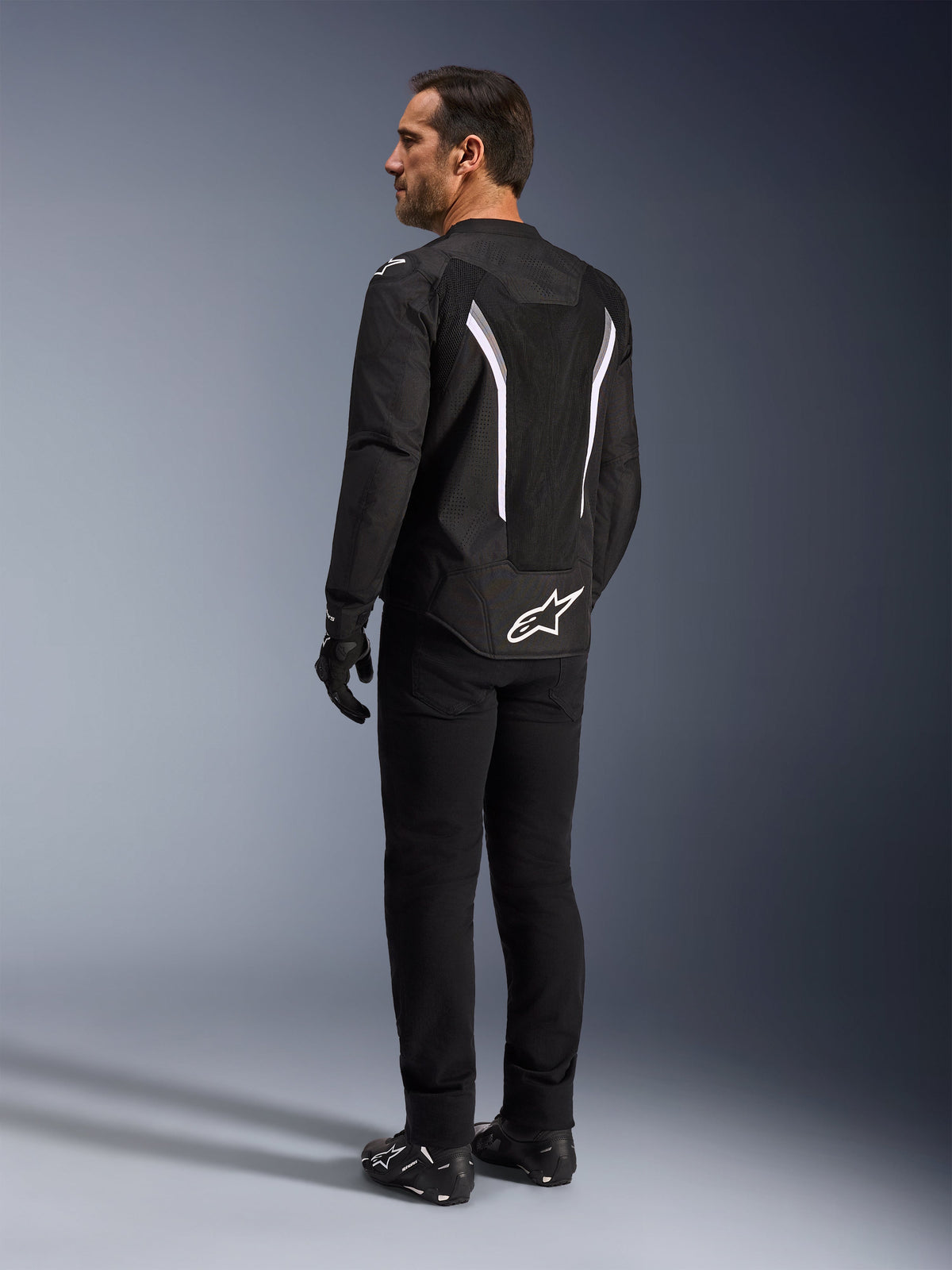 Alpinestars AST V3 Air Jacket, czarno-biała kurtka motocyklowa, widok z tyłu na modelu, z wstawkami z siatki zapewniającymi przepływ powietrza i białym logo na dole pleców, noszona z czarną techniczną odzieżą motocyklową.