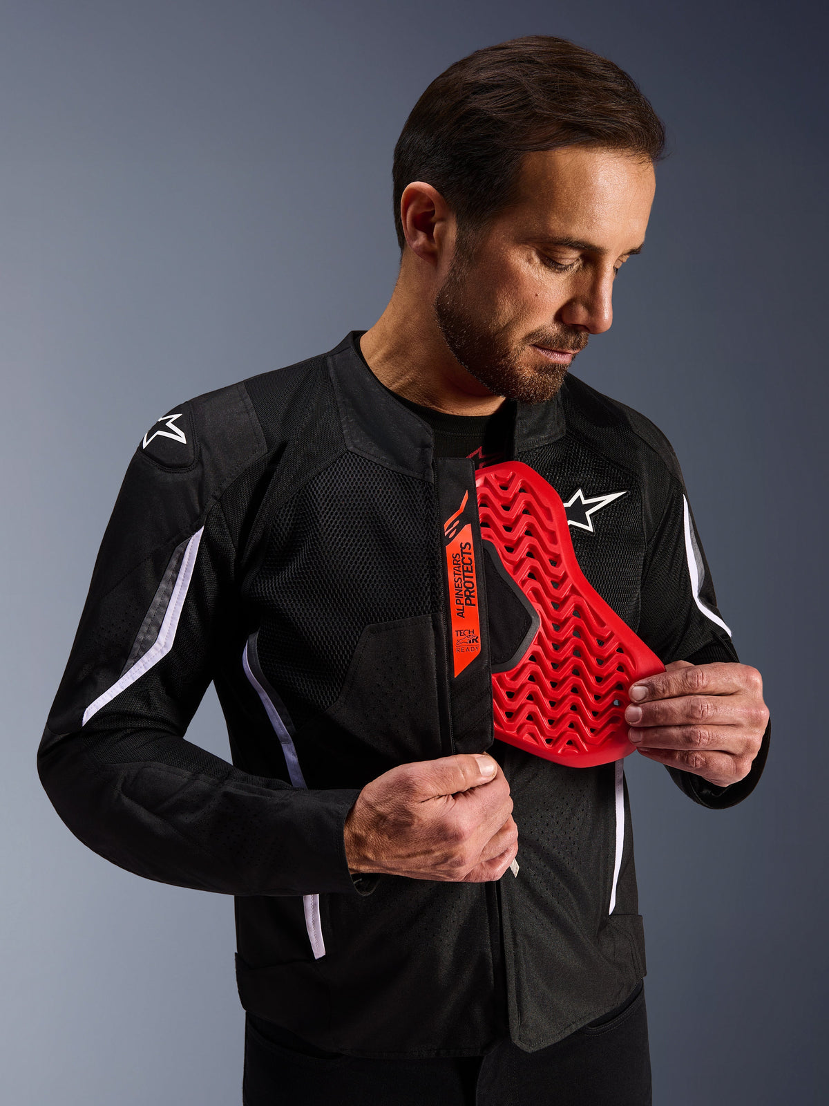 Alpinestars AST V3 Air Jacket w kolorze czarnym, mężczyzna odsłaniający jaskrawoczerwoną wkładkę ochronną klatki piersiowej o strukturze plastra miodu, podkreślająca kompatybilność z systemem Tech-Air Ready i zintegrowane ochraniacze dla bezpieczeństwa motocyklisty