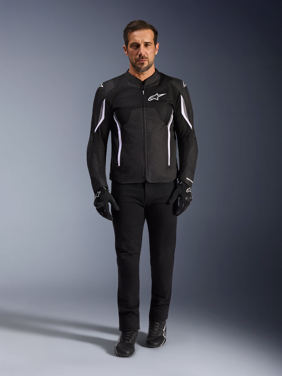 Męski model w kurtce Alpinestars AST V3 Air w kolorze czarnym z białymi akcentami, wyposażonej w panele wentylacyjne z siatki, o sportowym kroju, w zestawieniu z czarnymi technicznymi spodniami motocyklowymi, rękawicami i butami, widok z przodu, techniczna odzież motocyklowa do jazdy w ciepłe dni.
